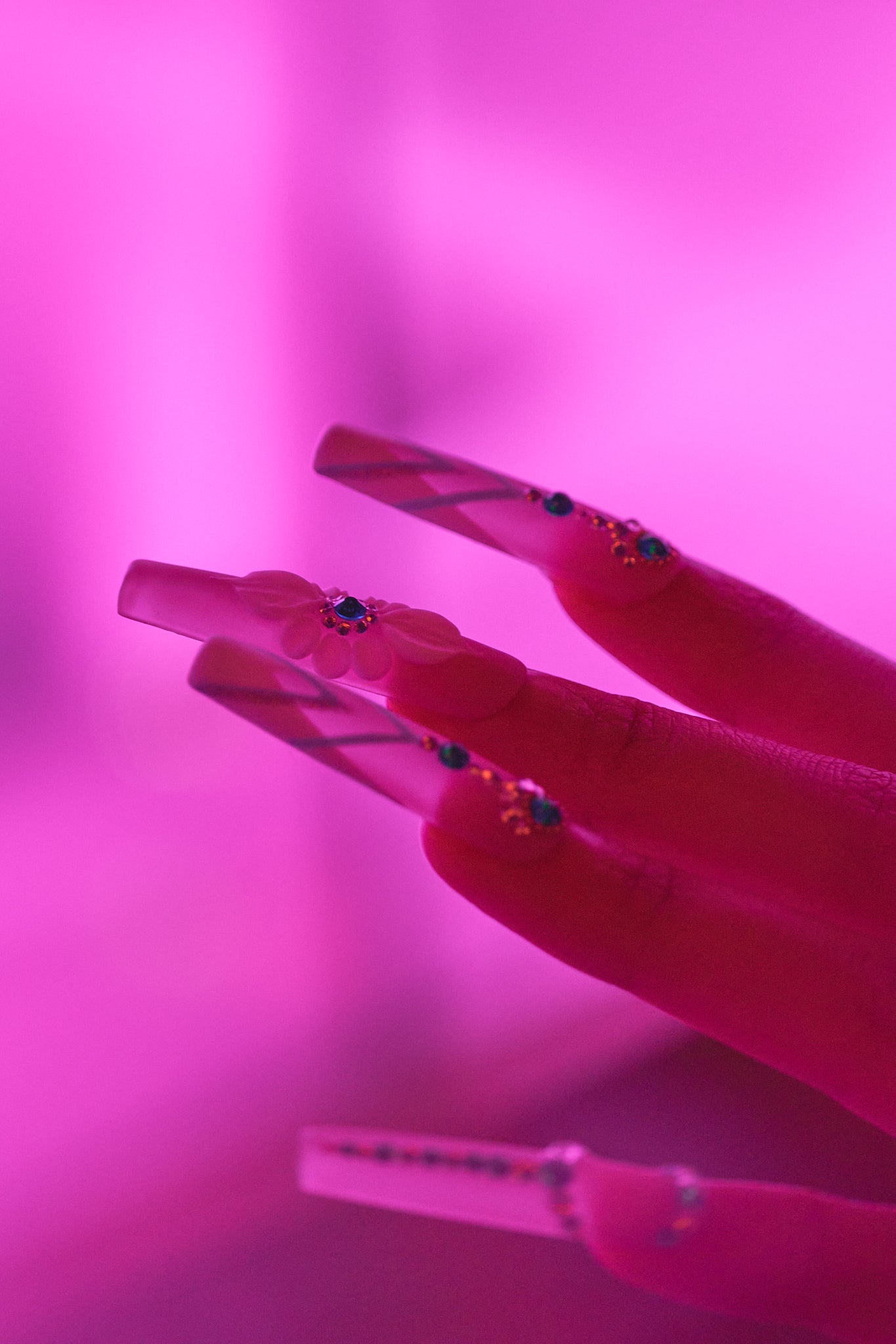 Backlit close-up of ‘Frost Waltz’ long square press-on nails with floral and rhinestone details against a soft pink glow.”
