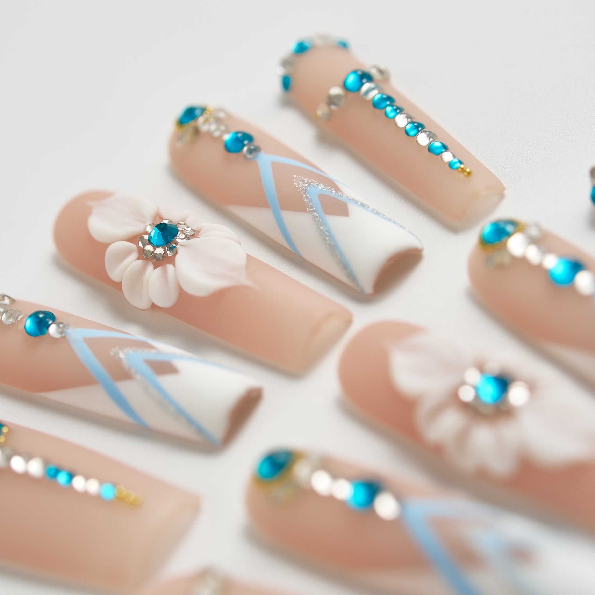 Macro shot of ‘Frost Waltz’ long square press-on nails with beige base, blue rhinestones, 3D floral accents, and silver-blue line detailing.
