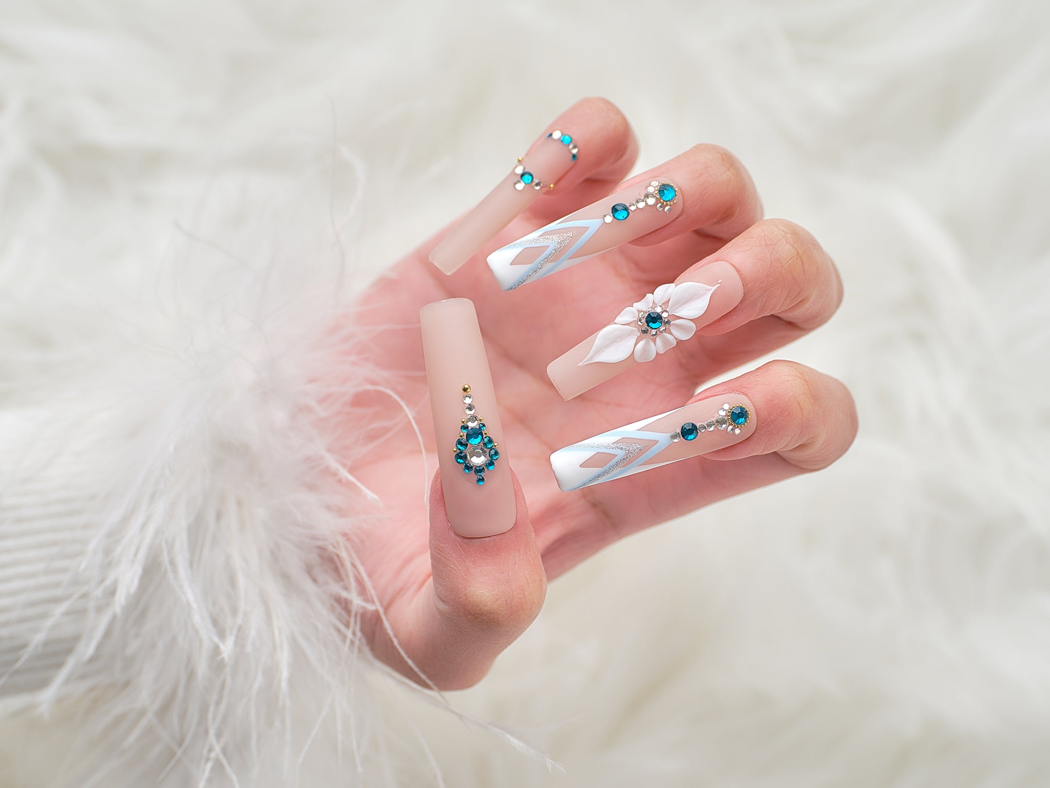 Hand wearing long square ‘Frost Waltz’ press-on nails with beige base, blue gems, and 3D floral accents, posed against soft white faux fur.