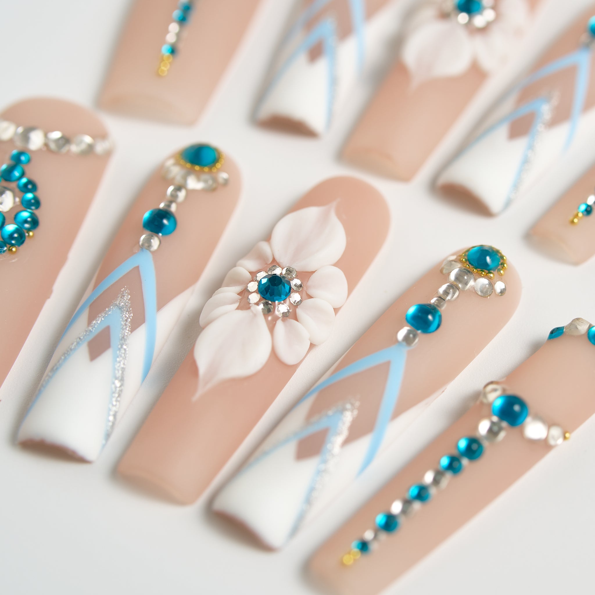 Top-down macro of ‘Frost Waltz’ long square press-on nails, featuring white 3D floral art, blue rhinestones, and geometric white-blue patterns on a beige base.