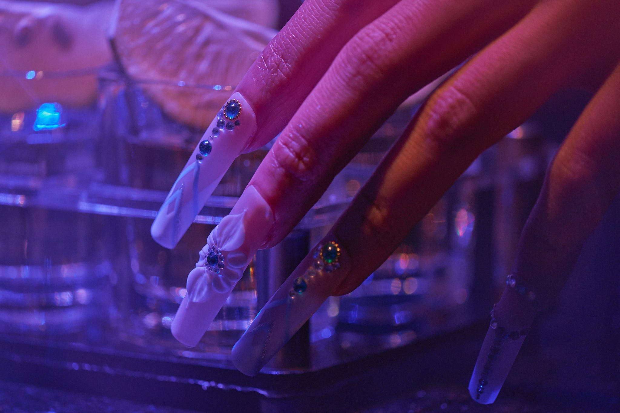 Close-up of hand wearing ‘Frost Waltz’ long square press-on nails with crystal and floral accents, lit by violet neon light in a nightlife bar setting.”