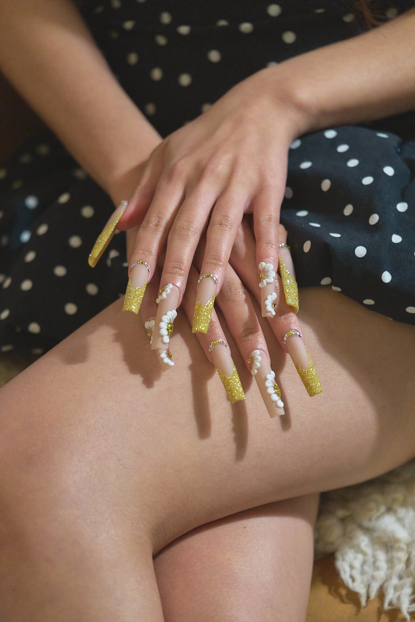 Hands with MiuNail “Golden Bloom” long square glitter yellow press-on nails featuring 3D pearl embellishments, styled on crossed legs in a dotted dress.