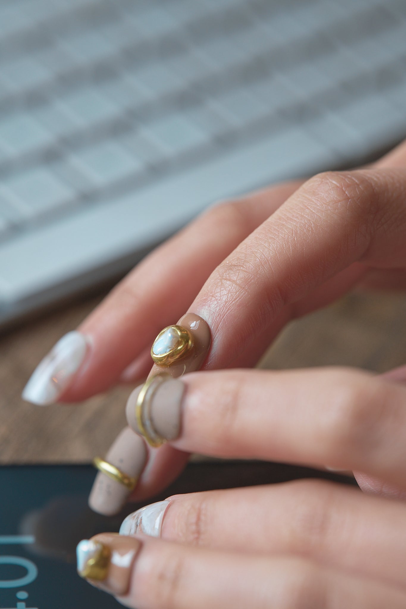 Close-up of a hand with MiuNail Golden Dune press-on nails in marble, brown, and gold details, holding a phone with a wireless keyboard blurred in the background.