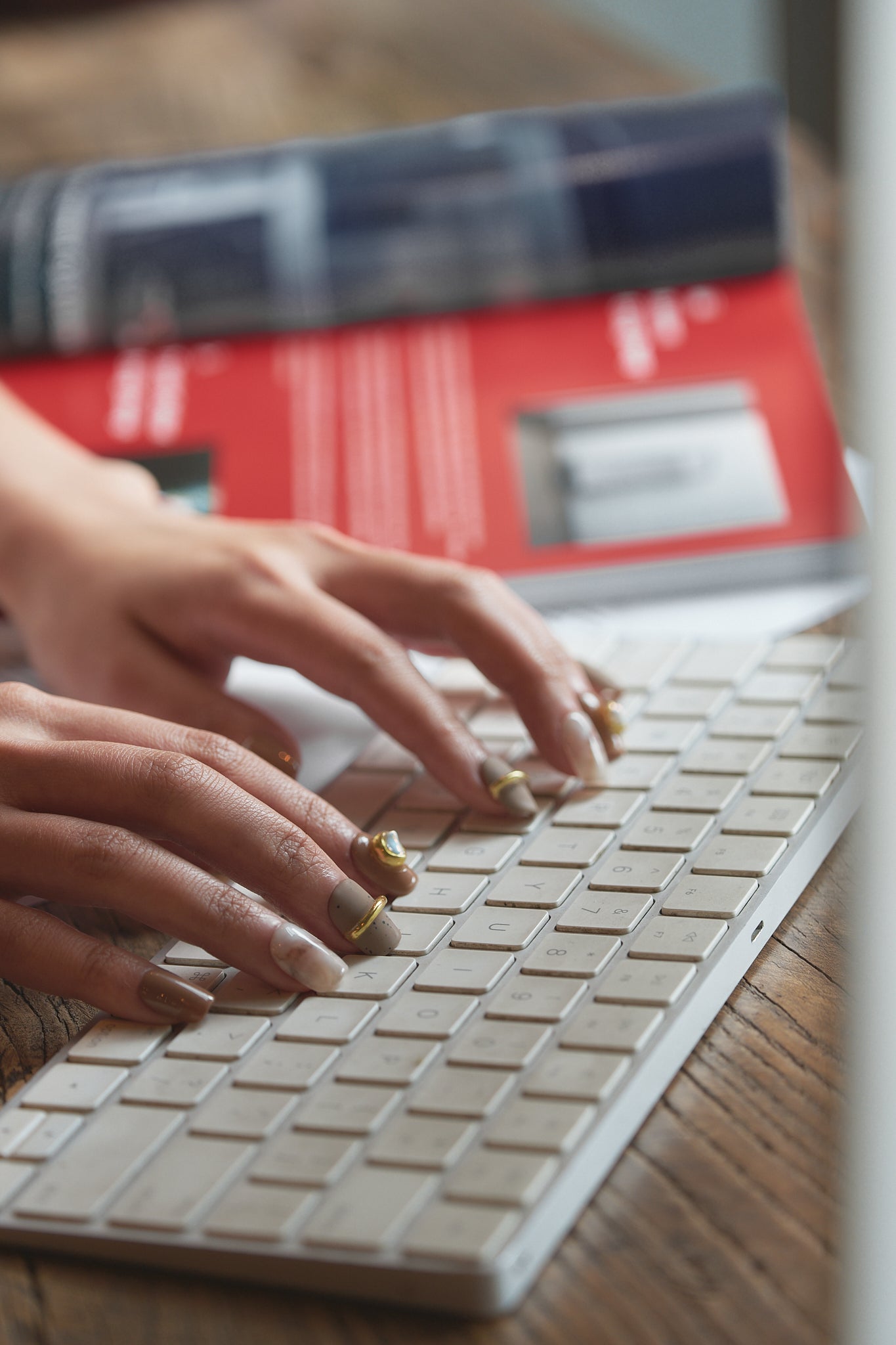 Hands with MiuNail Golden Dune press-on nails in marble, brown, and gold, typing on a white wireless keyboard with a red book and magazine in the background.