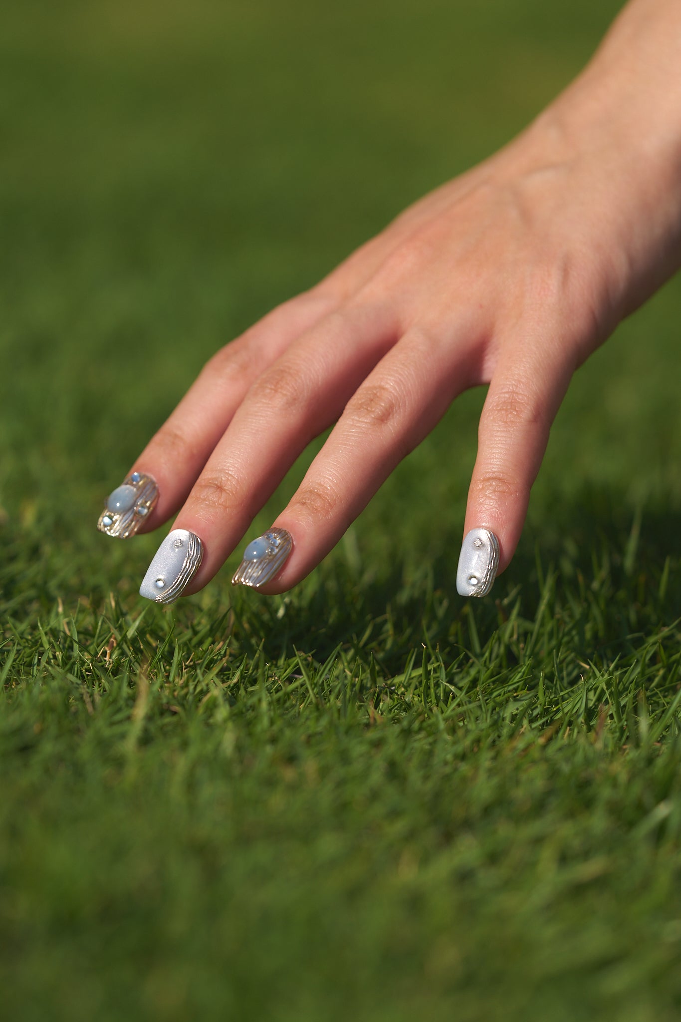 Hand with MiuNail Mist Orbit gray and silver press-on nails with rhinestone details, gently placed on green grass in natural sunlight.