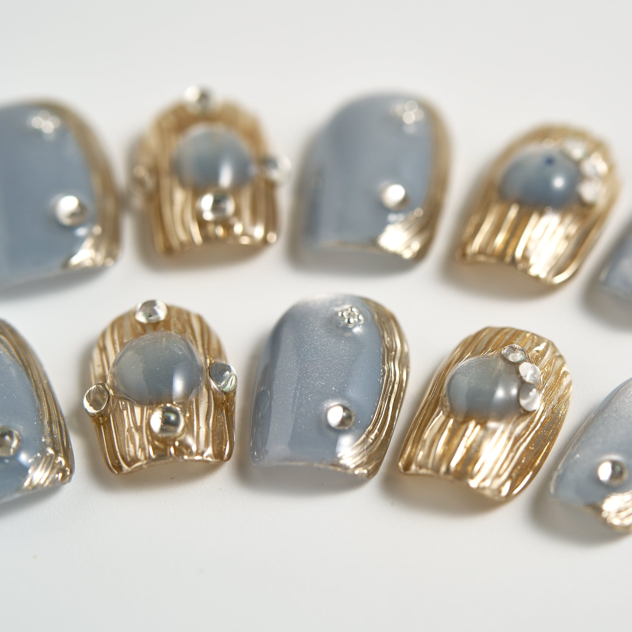 Detailed view of MiuNail Mist Orbit press-on toenails with metallic gray and gold textures, arranged on a light background.