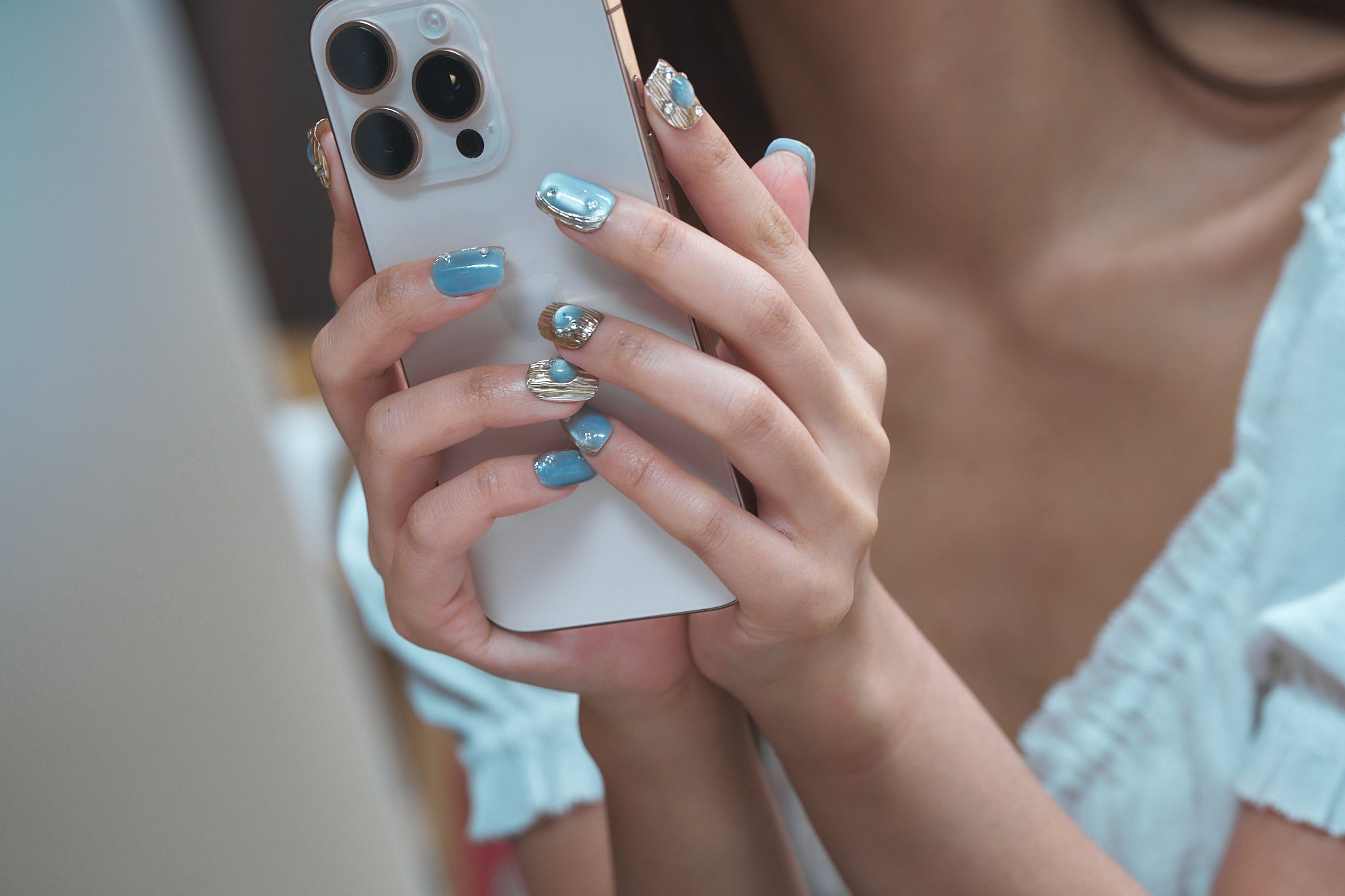 Hands with MiuNail Mist Orbit press-on nails in metallic silver and gold, gently holding a smartphone.