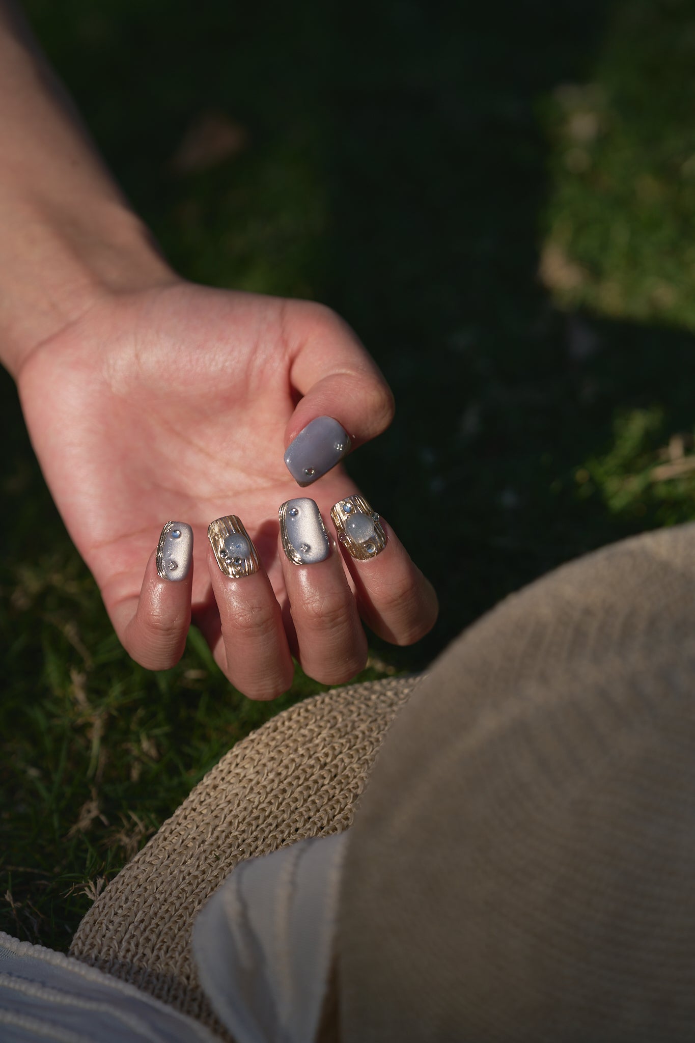 Hand with MiuNail Mist Orbit press-on nails shown in natural sunlight, resting on a straw hat outdoors.