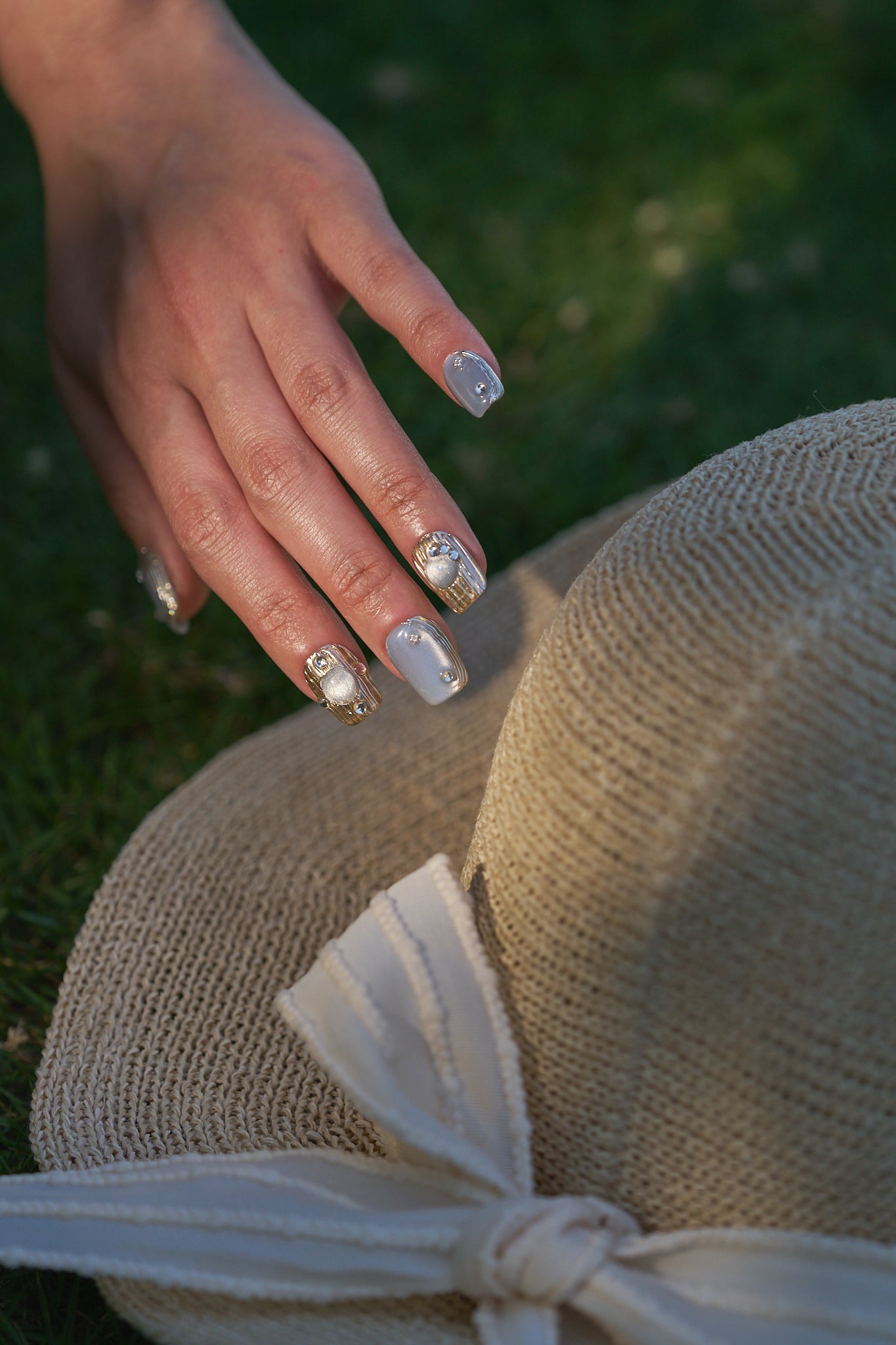 Hand showing MiuNail Mist Orbit press-on nails, resting on a straw hat with a soft outdoor background.