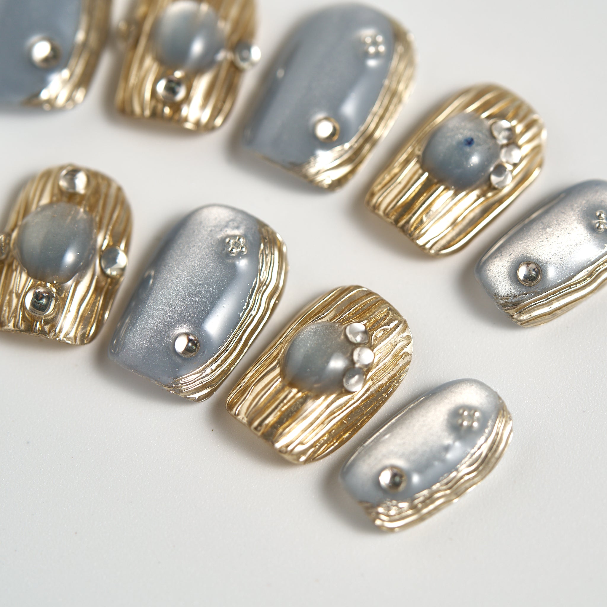 Close-up of MiuNail Mist Orbit press-on toe nails featuring metallic silver and gold textured accents, shown on a white surface.