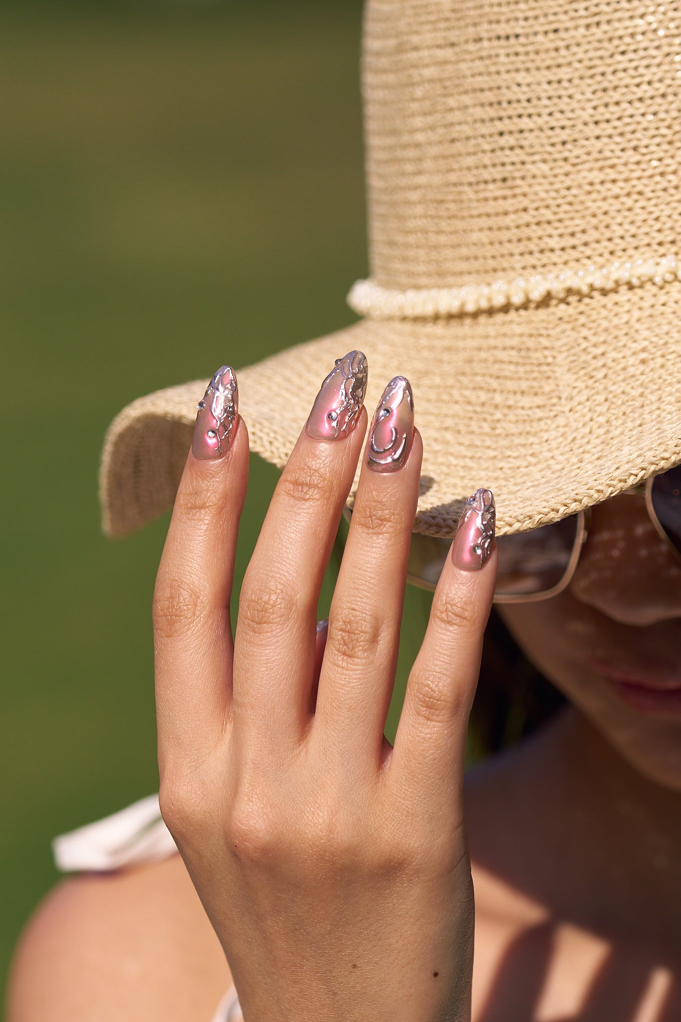 Hand wearing MiuNail Molten Coral press-on nails in coral pink with silver chrome swirls and a raised floral charm, fingers resting on a bag strap.