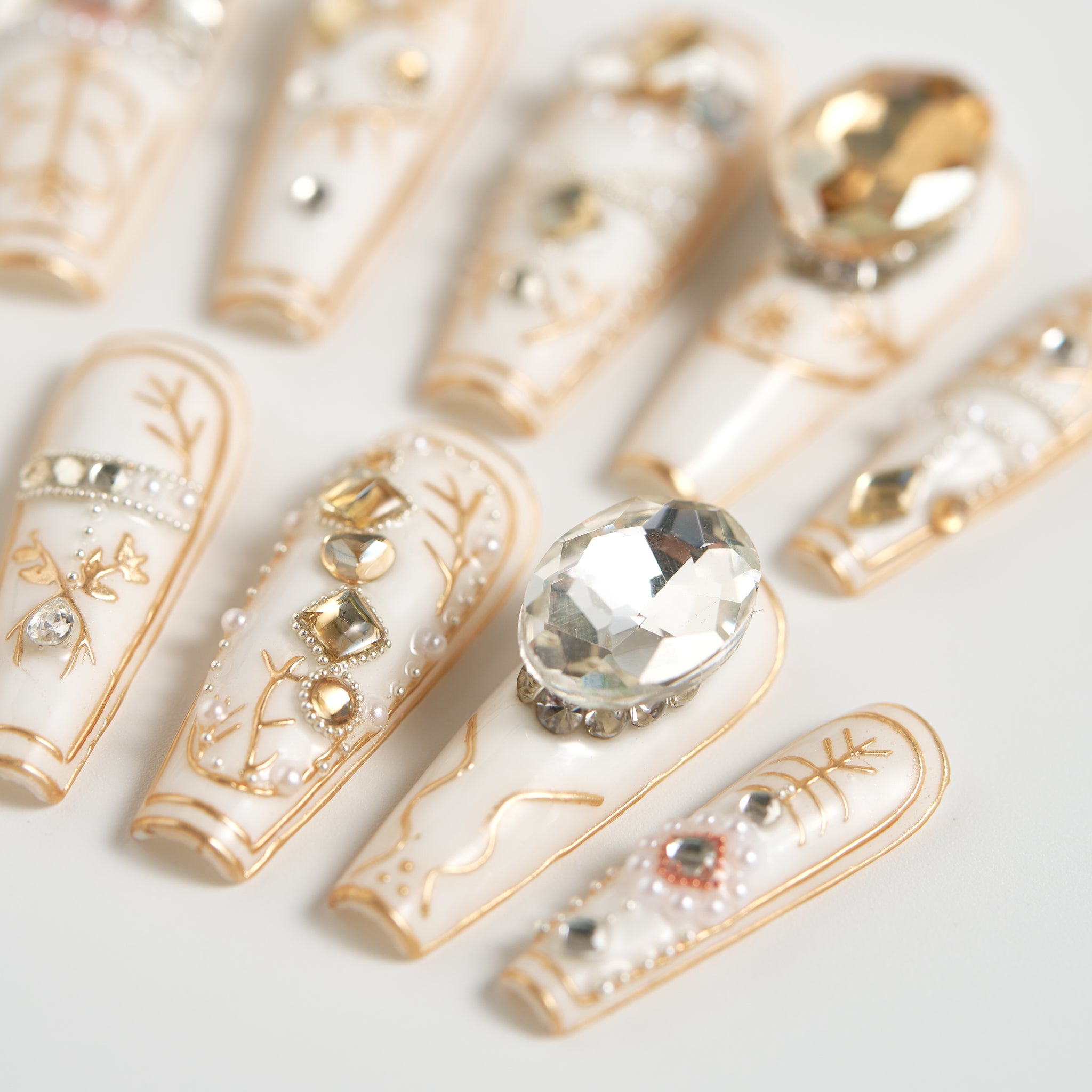 Close-up of MiuNail Ornate Veil press-on nails with white base, gold line designs, and oversized clear crystal accents, arranged on a white background.