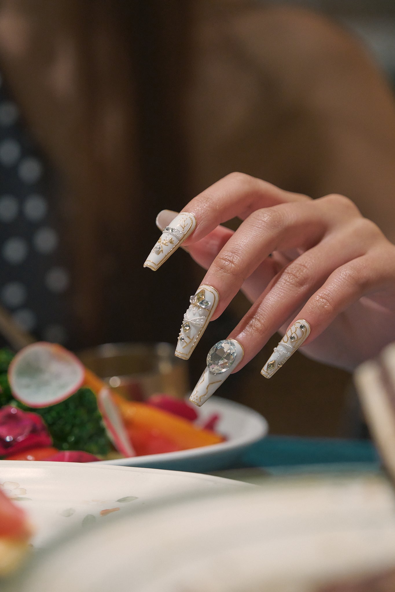 Hand with detailed white and gold nail art resting above a colorful salad at a dinner table.