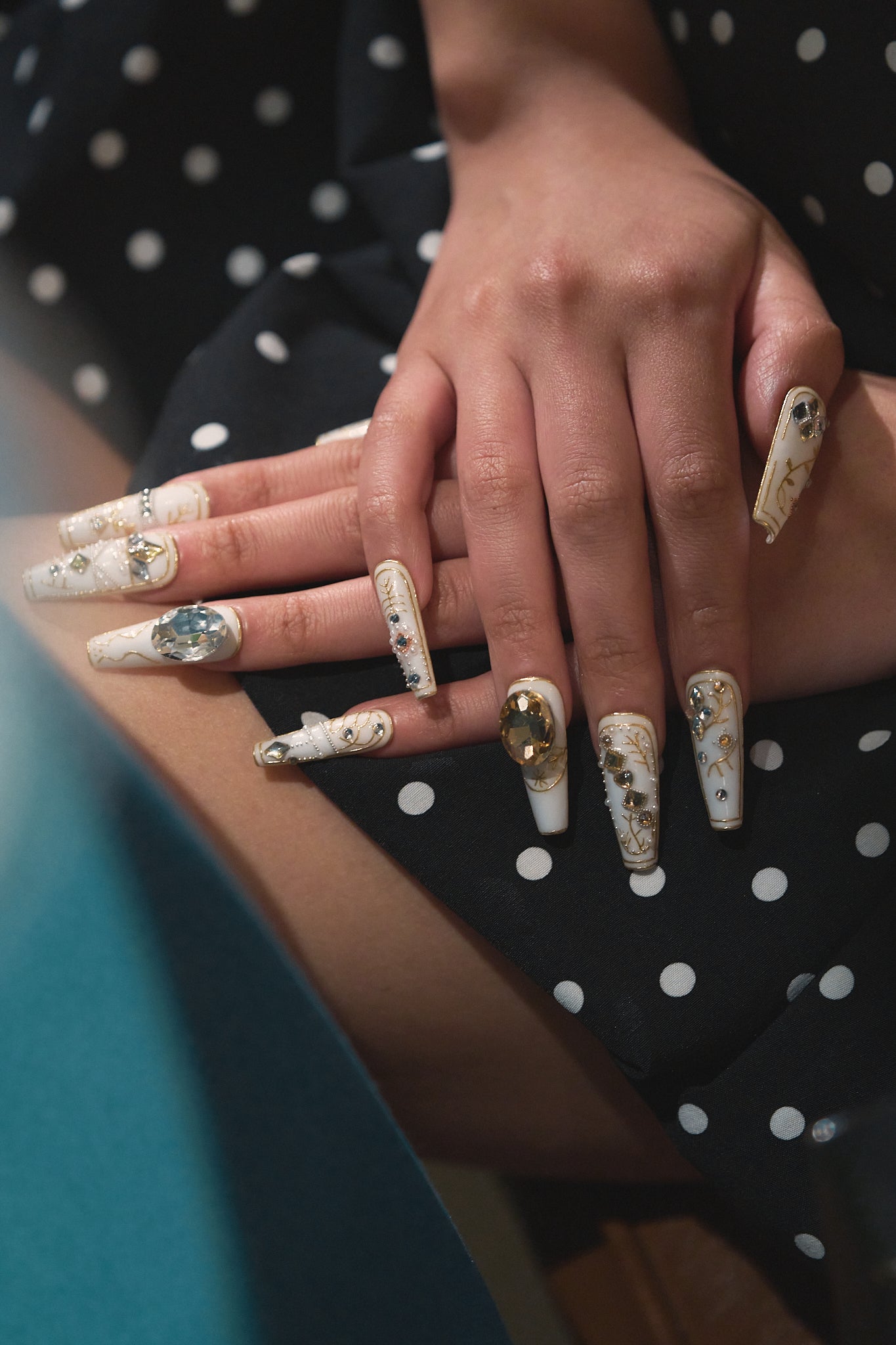 Both hands with gold-accented white nail art resting on a black polka dot dress, showing the complete manicure look.