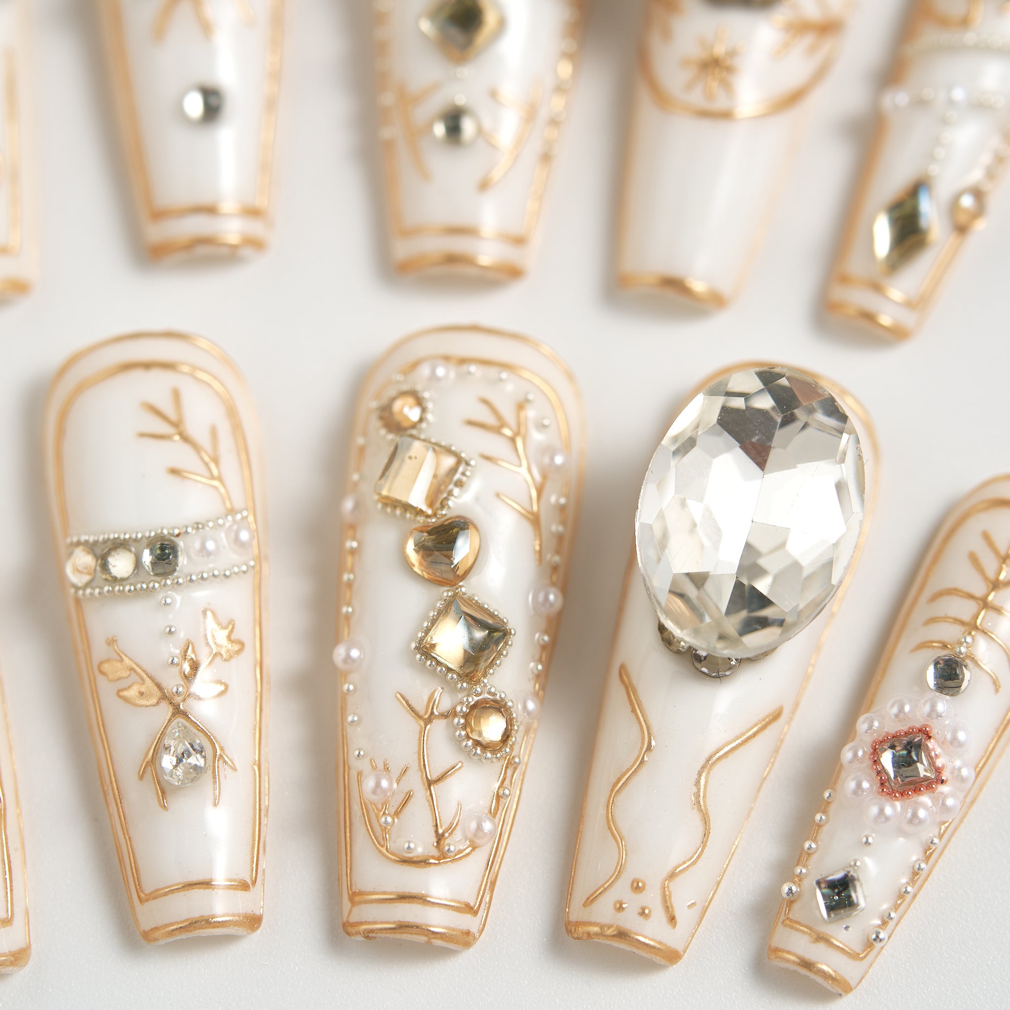 Close-up of MiuNail Ornate Veil press-on nails in glossy white with gold line art and large crystal embellishments, arranged on a white background.