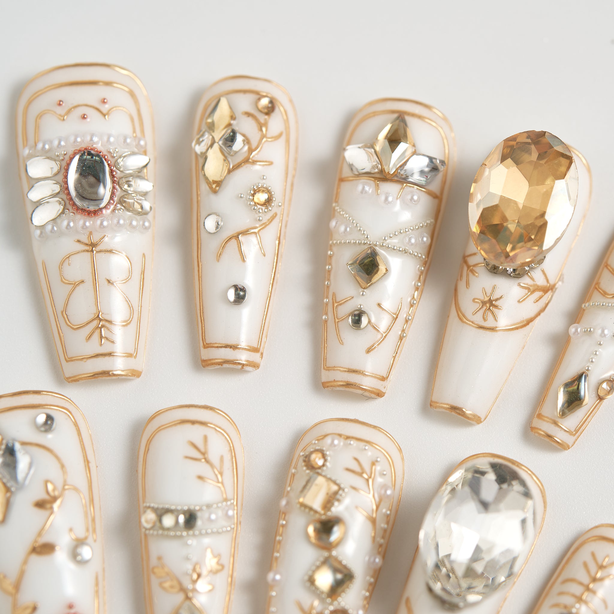 MiuNail Ornate Veil press-on nails in glossy white with gold line art, silver and gold crystal embellishments, detailed close-up on a white background.