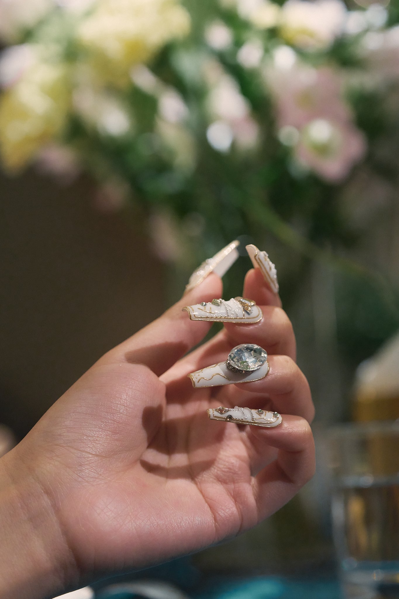 Hand with intricate white and gold nails, holding fingers slightly curved in front of a softly blurred floral background.