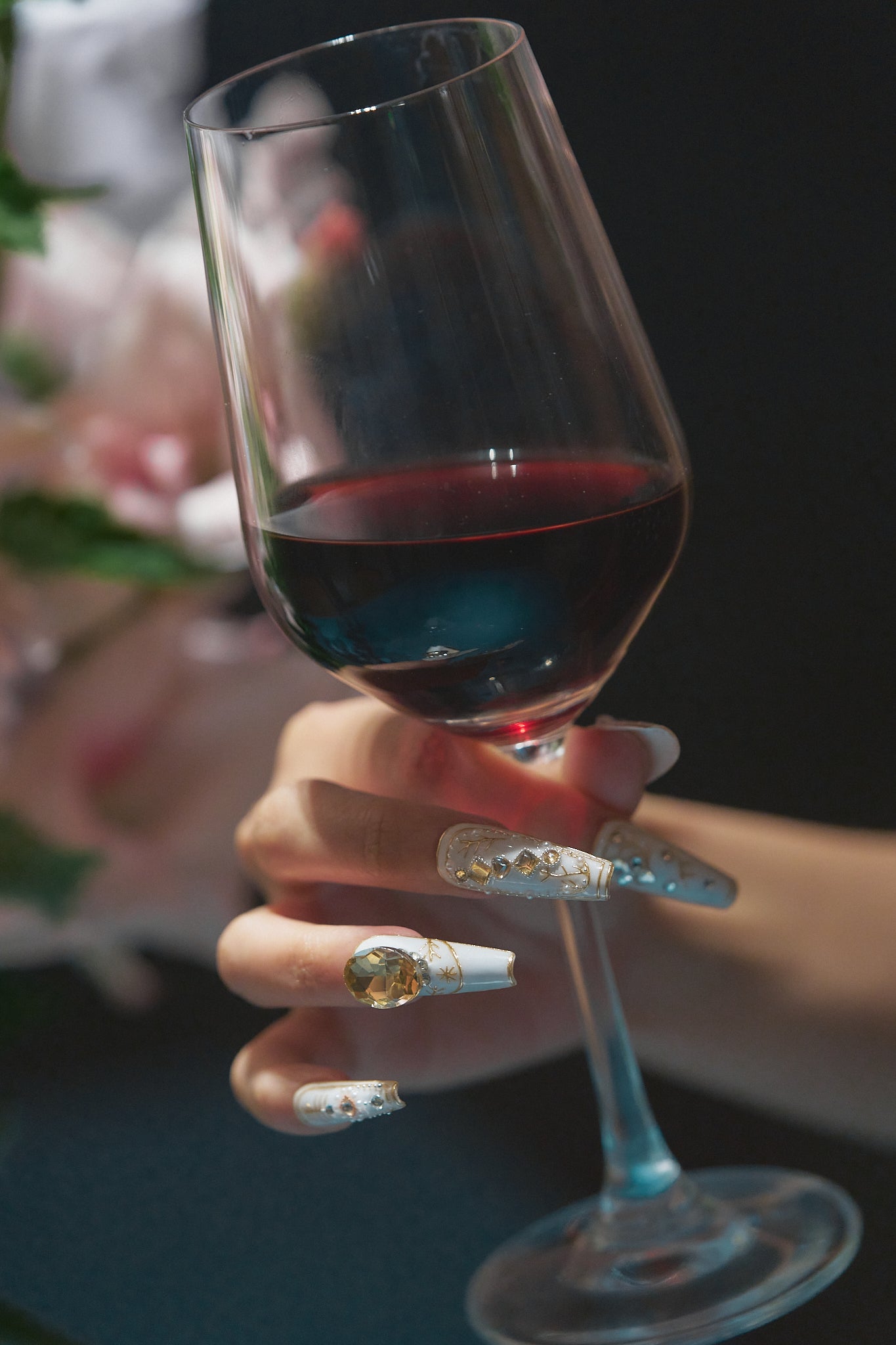 Hand with elegant white and gold embellished nails gently holding a glass of red wine, soft floral background out of focus.