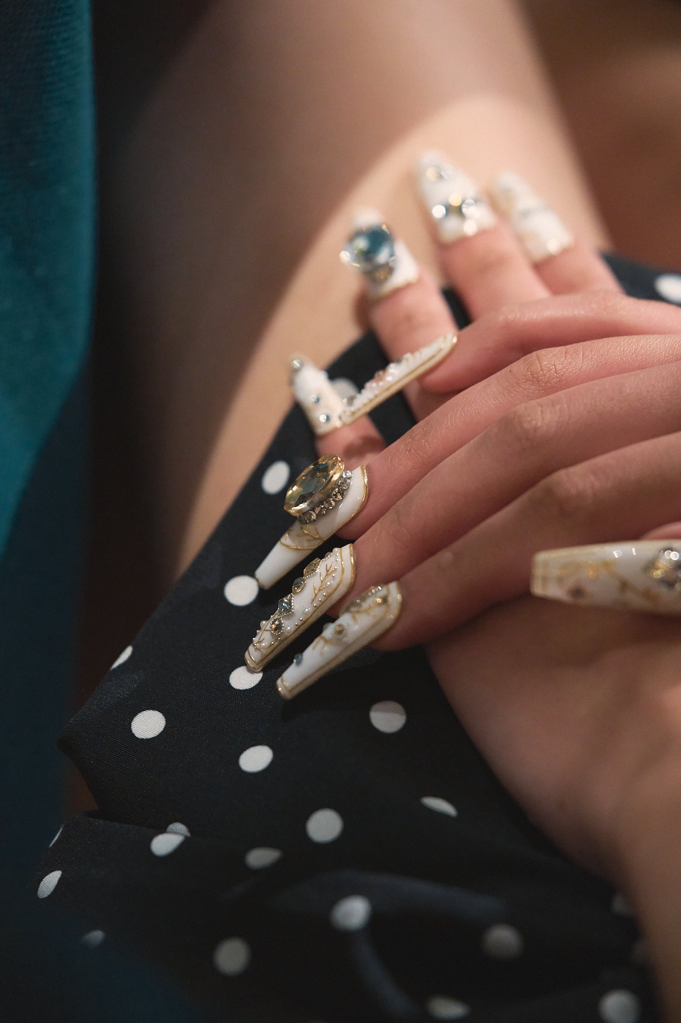 Hand with detailed gold and white nail designs and statement rings, resting on a black polka dot fabric.