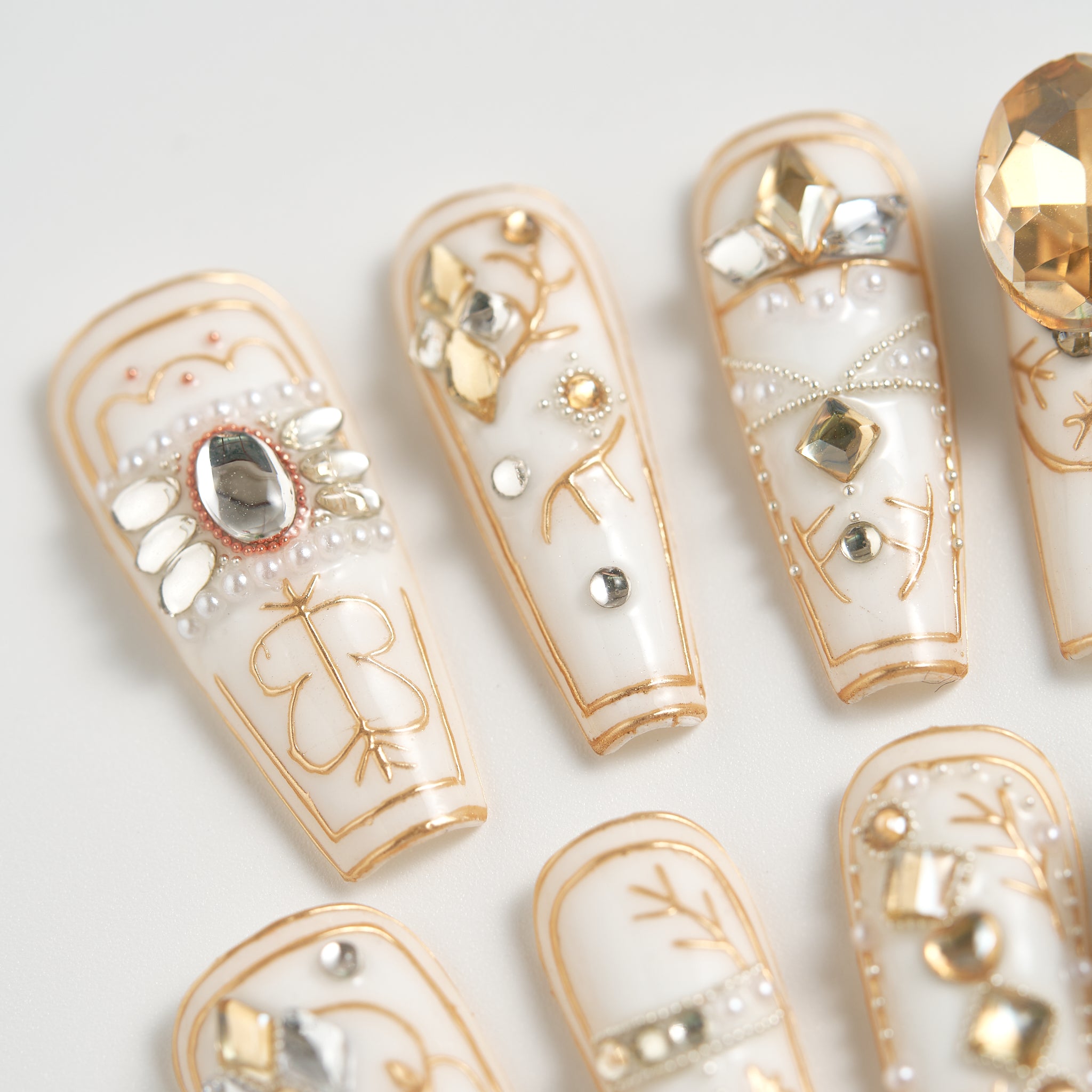 Close-up of MiuNail Ornate Veil press-on nails featuring gold and silver embellishments, hand-drawn filigree, and pearl details on a white background.