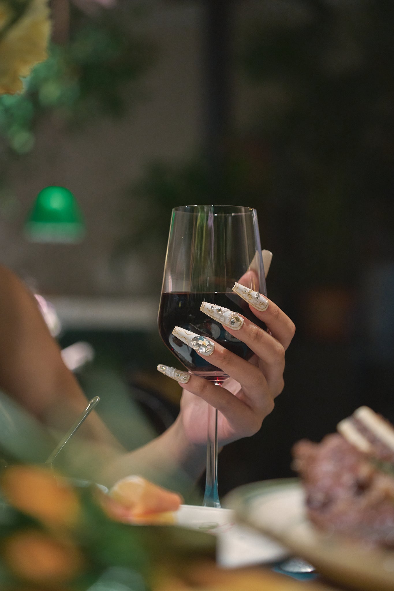 Hand wearing MiuNail Ornate Veil press-on nails with gold and crystal embellishments, elegantly holding a glass of red wine at a dinner table.