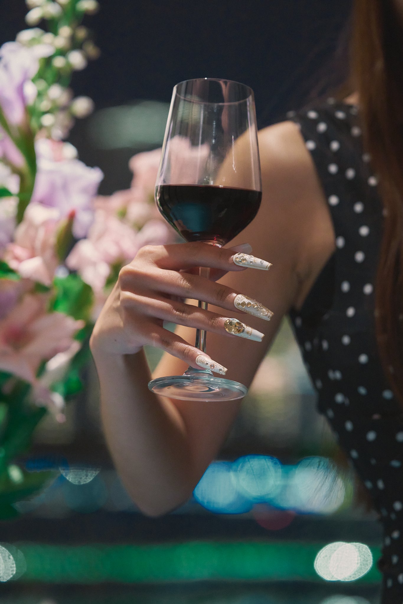 Hand wearing MiuNail Ornate Veil press-on nails with gold and crystal details, holding a glass of red wine with blurred flowers and lights in the background.