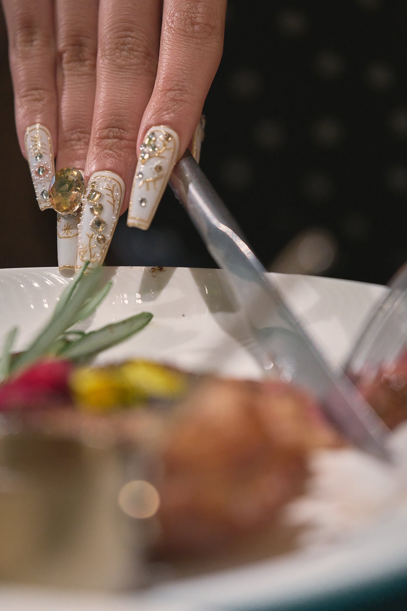 Hand with ornate nail designs using tongs to serve food, focus on nail art details and dining scene.