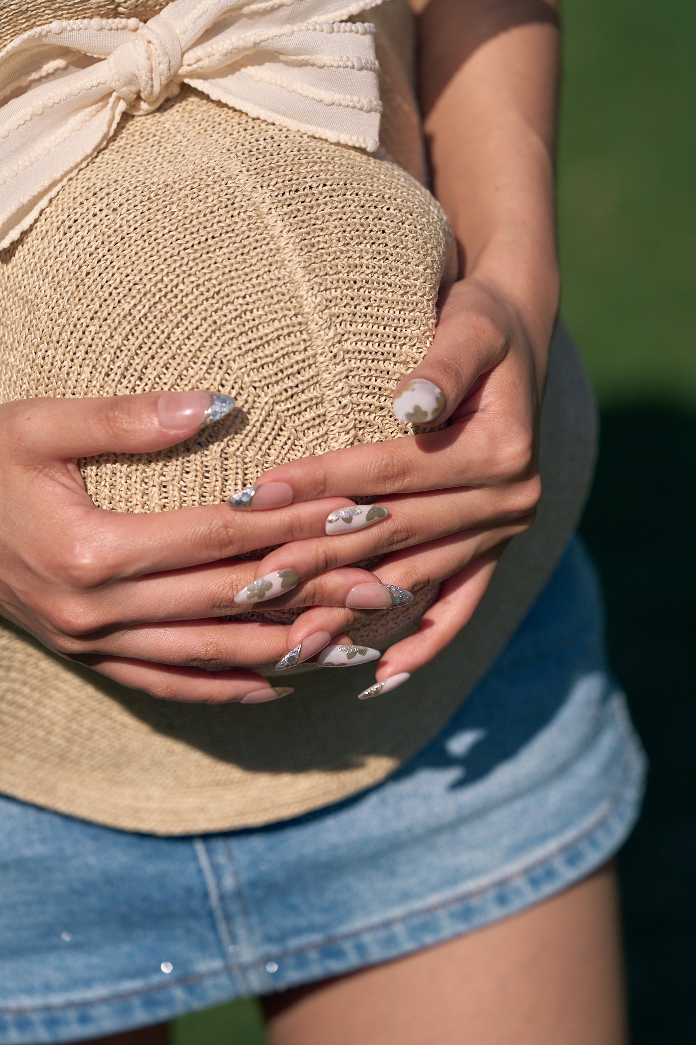 Petal Bloom long almond press-on nails in marble white and gold, hands gently holding a straw hat over a maternity belly outdoors in denim skirt