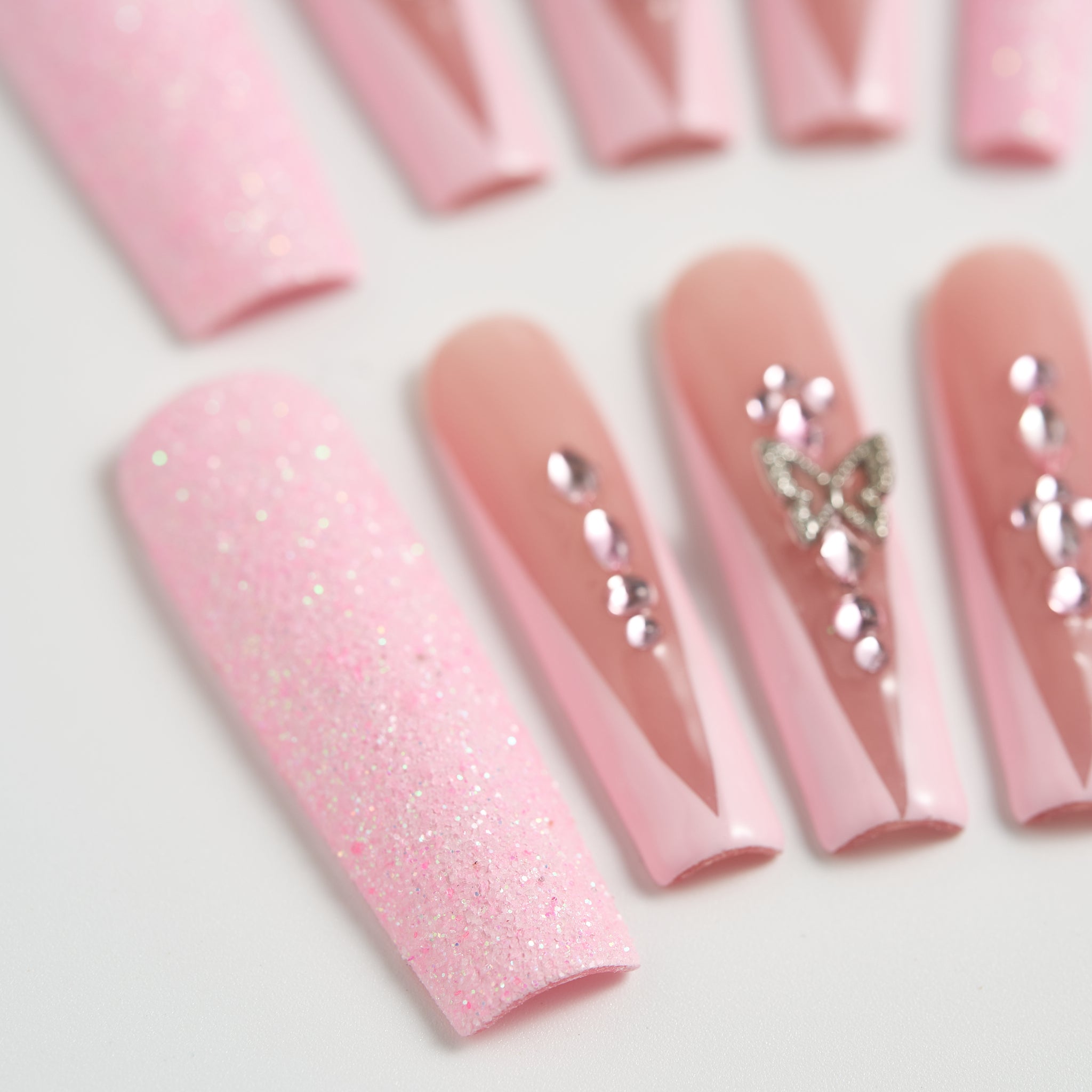 Close-up of Sugar Flutter press-on nails showing the transition from pastel pink glitter to glossy nude, detailed with silver rhinestones and a 3D butterfly charm, on a white background.