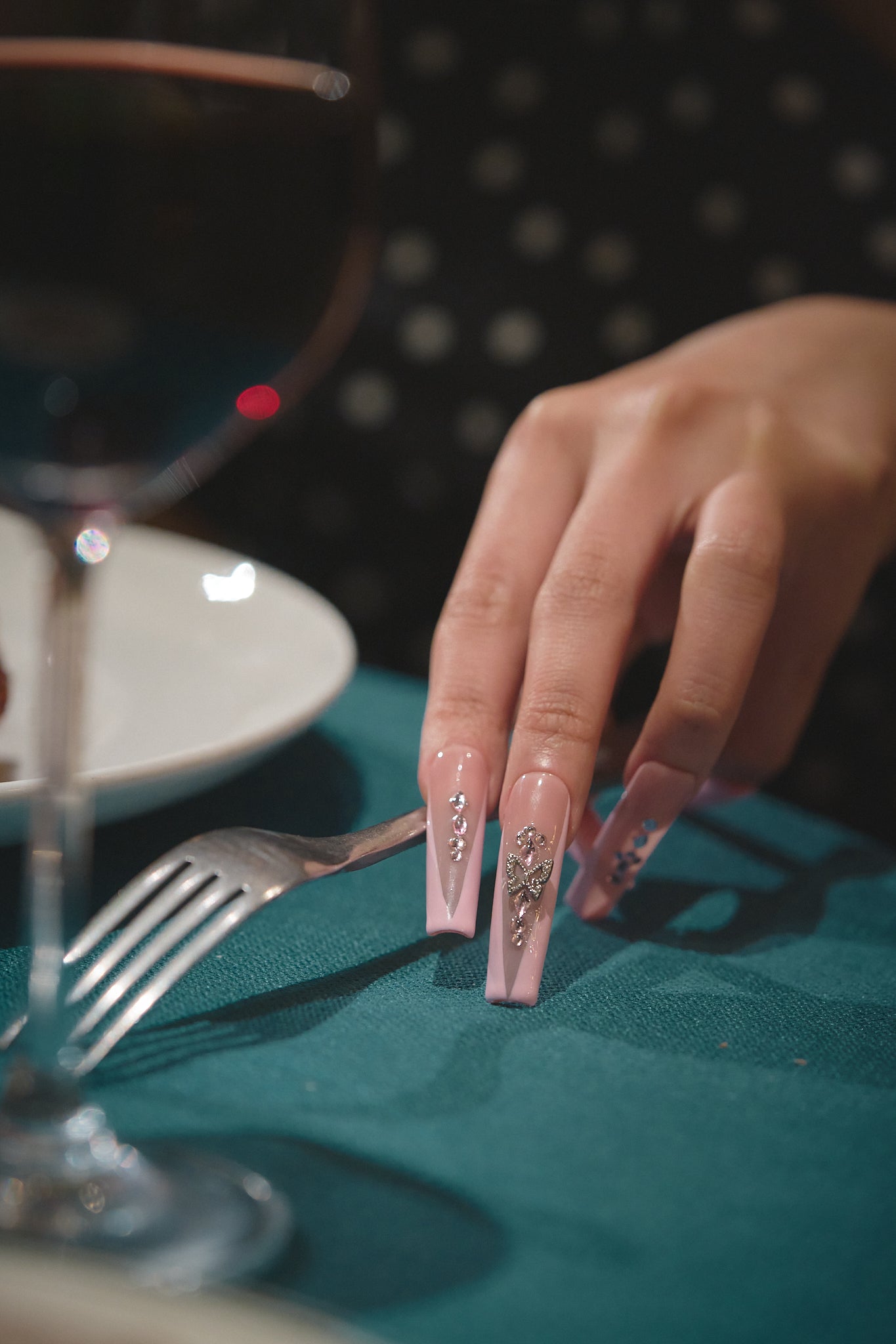 Hand with soft pink and crystal-embellished nails resting on a teal tablecloth, holding a fork beside a glass of wine at a dinner setting.