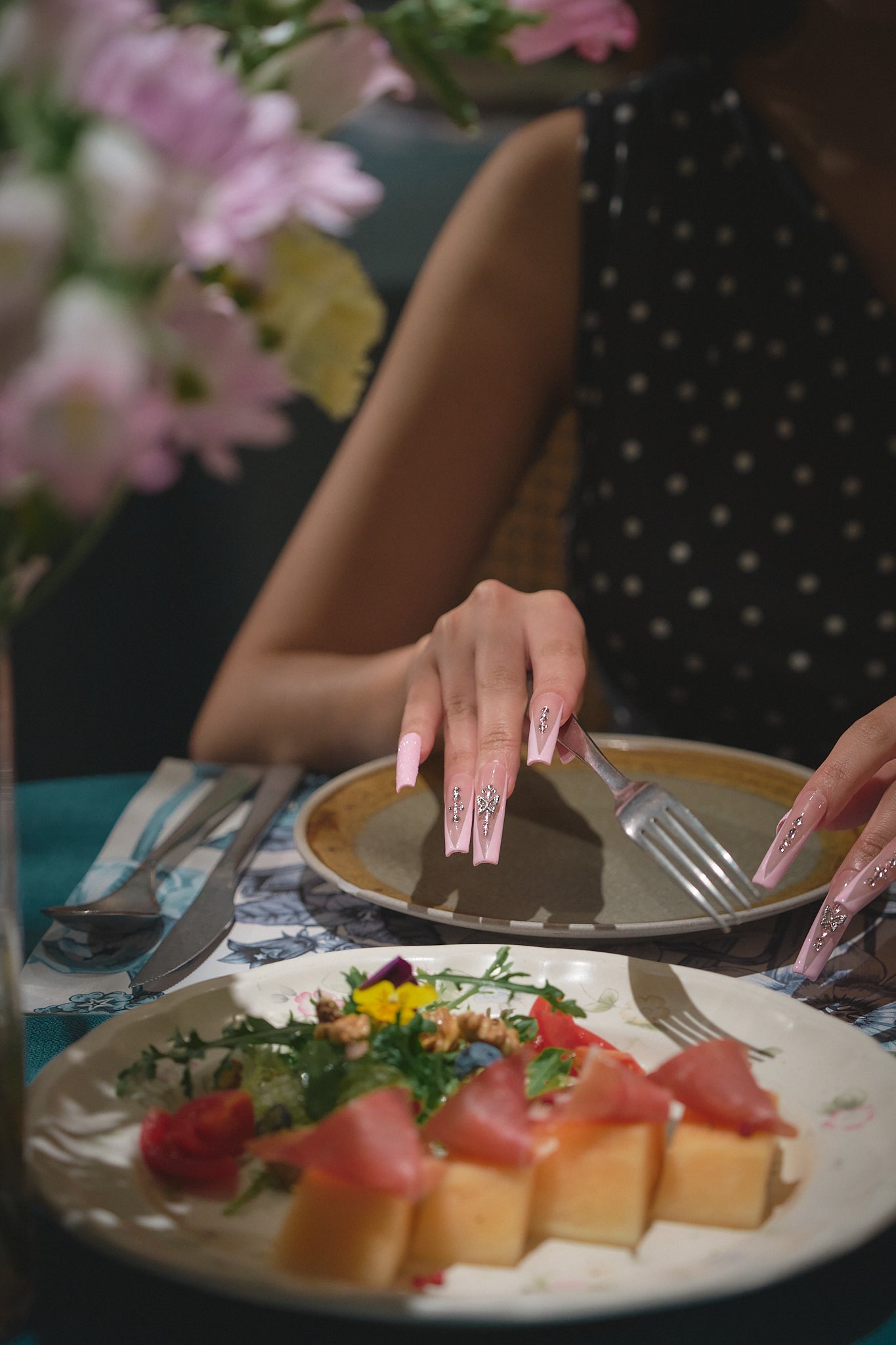 Woman with long pink glitter nails using fork and knife at a dinner table, surrounded by flowers and elegant place settings.