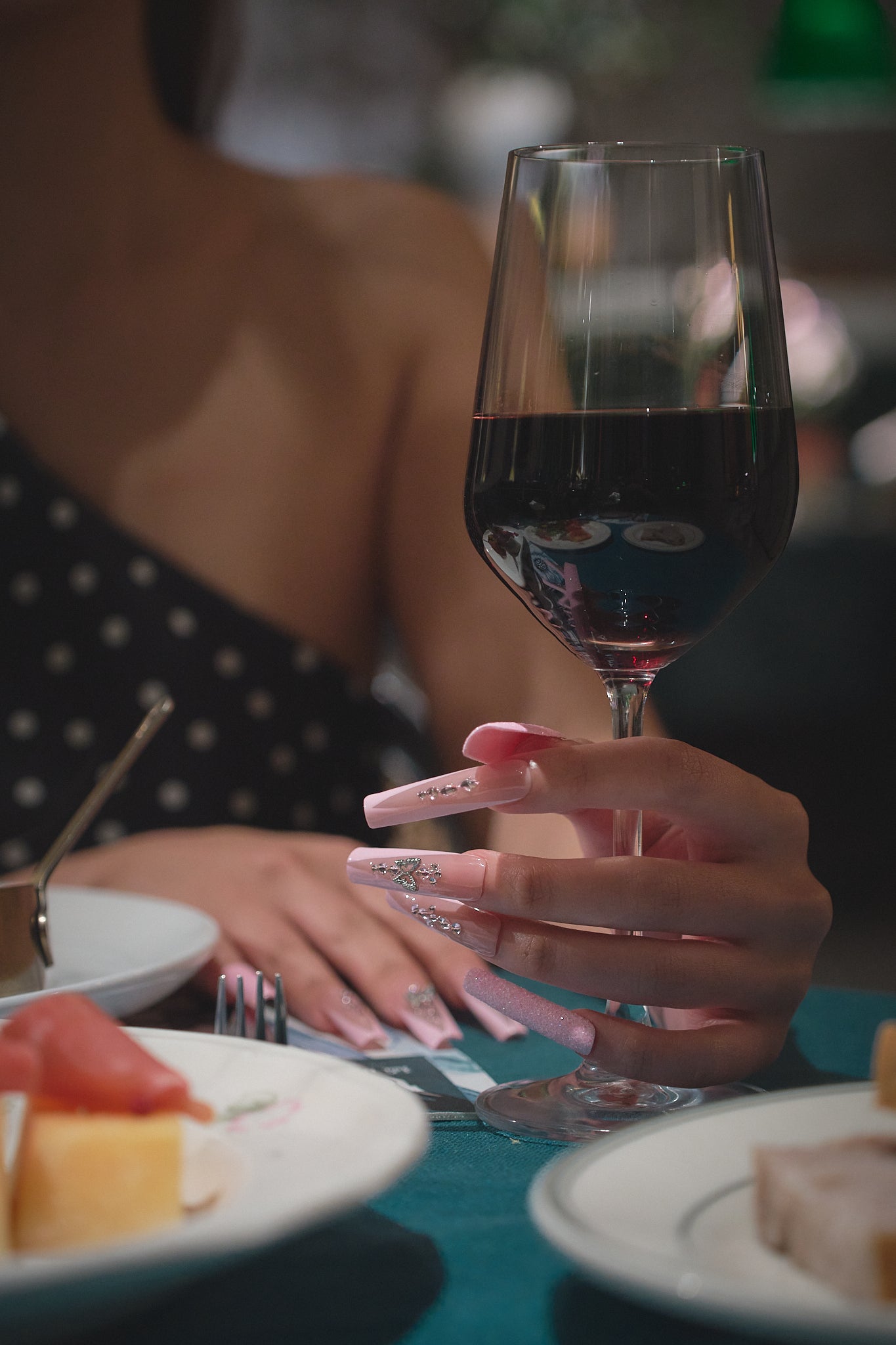 Woman’s hand with pink crystal-embellished nails gently holding a glass of red wine at a dinner table, with plates of food and an elegant setting in the background.