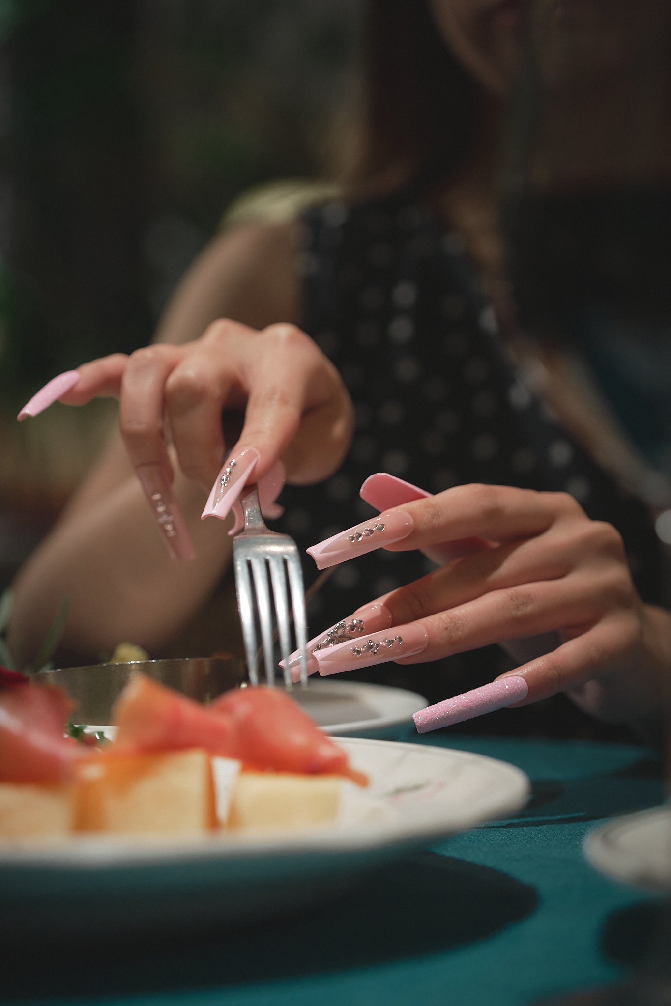 Hands with long, sparkly pink nails and rhinestone details holding a fork and knife above a plate of fruit; elegant dinner setting, soft background.