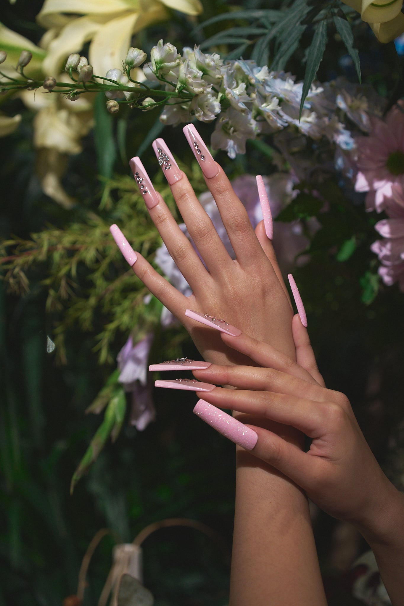 Two hands with extra-long pastel pink nails and rhinestone accents posed gracefully in front of blooming flowers.