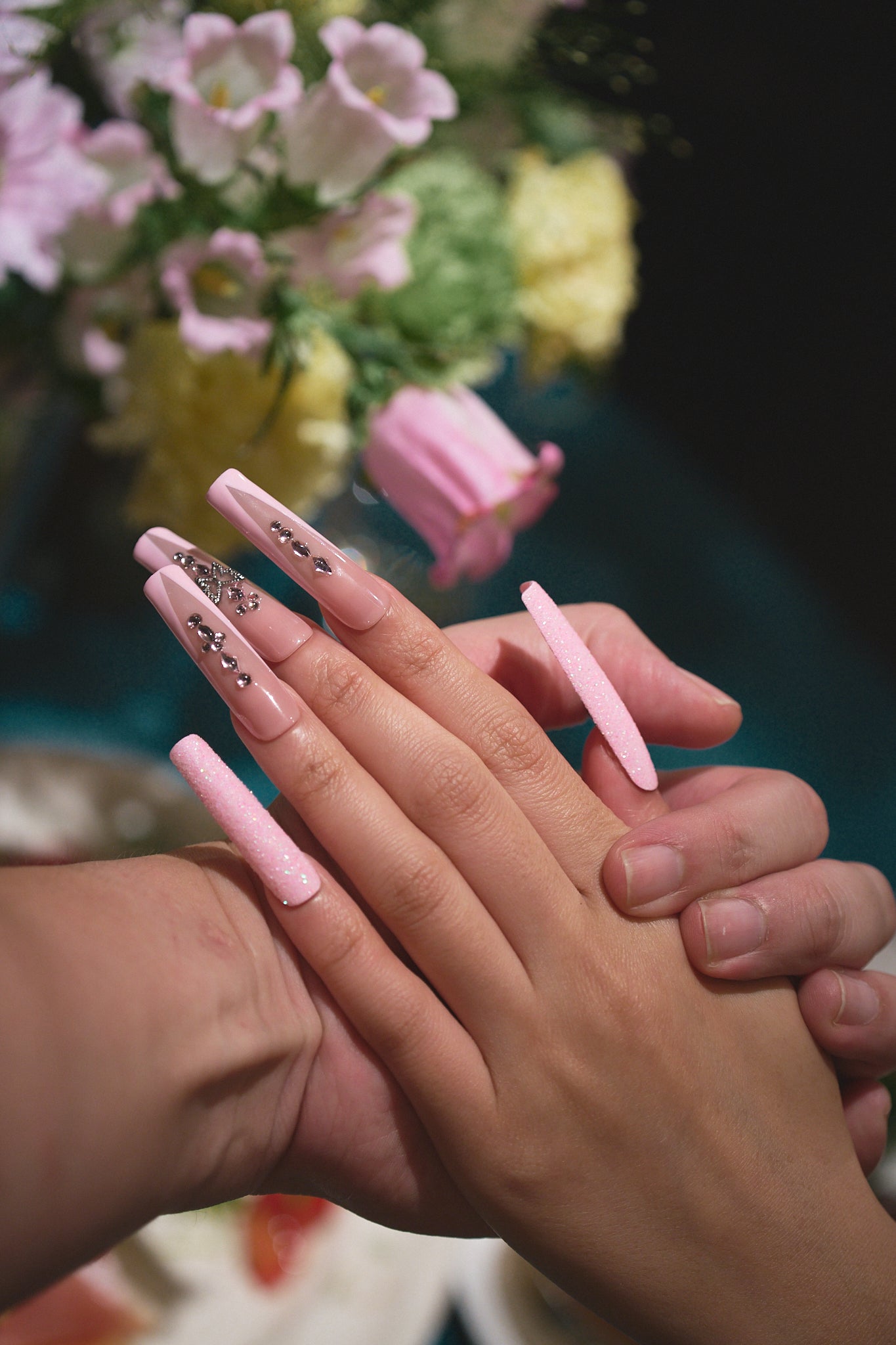 A gentle hand hold, featuring long pink glitter nails with rhinestone accents; blurred floral bouquet in the background, soft and romantic atmosphere.