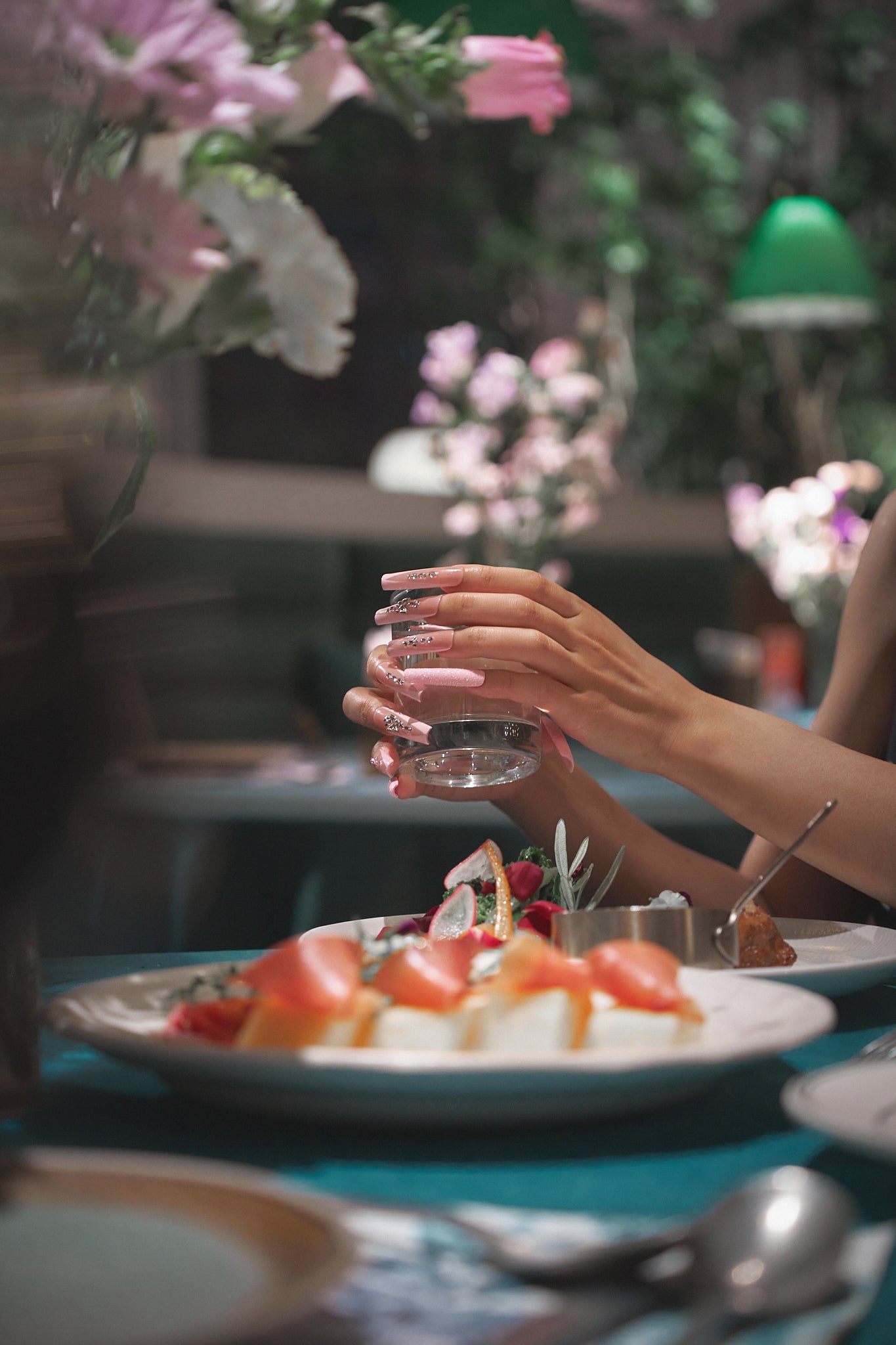 Elegant hands with long, pastel pink nails gently holding a glass of water at a beautifully set dining table; lush florals and gourmet dishes in the softly blurred background.