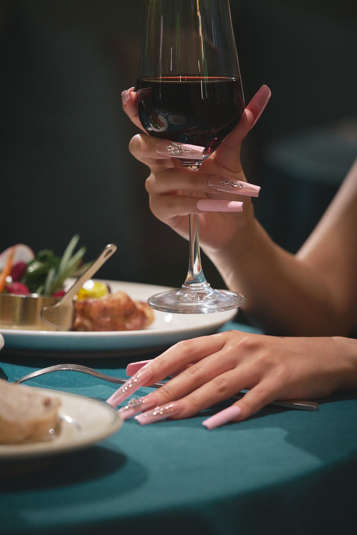 A graceful hand with glossy pastel pink nails holds a glass of red wine above a teal tablecloth, with elegant plating and soft background blur.