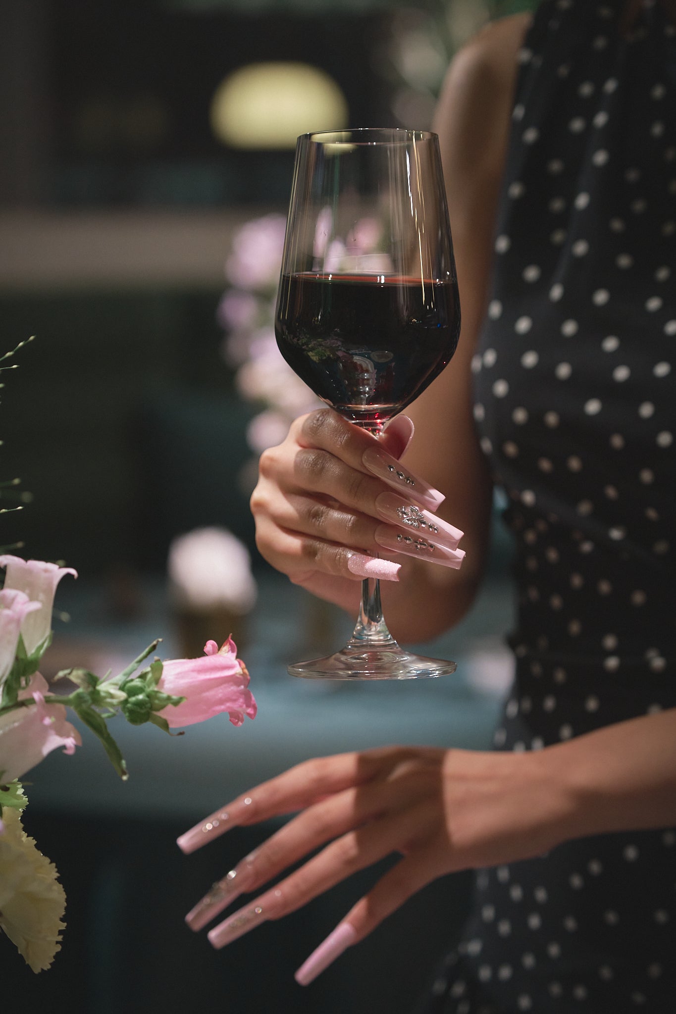 Woman in a polka dot dress holding a glass of red wine, showing Sugar Flutter pink press-on nails by MiuNail.
