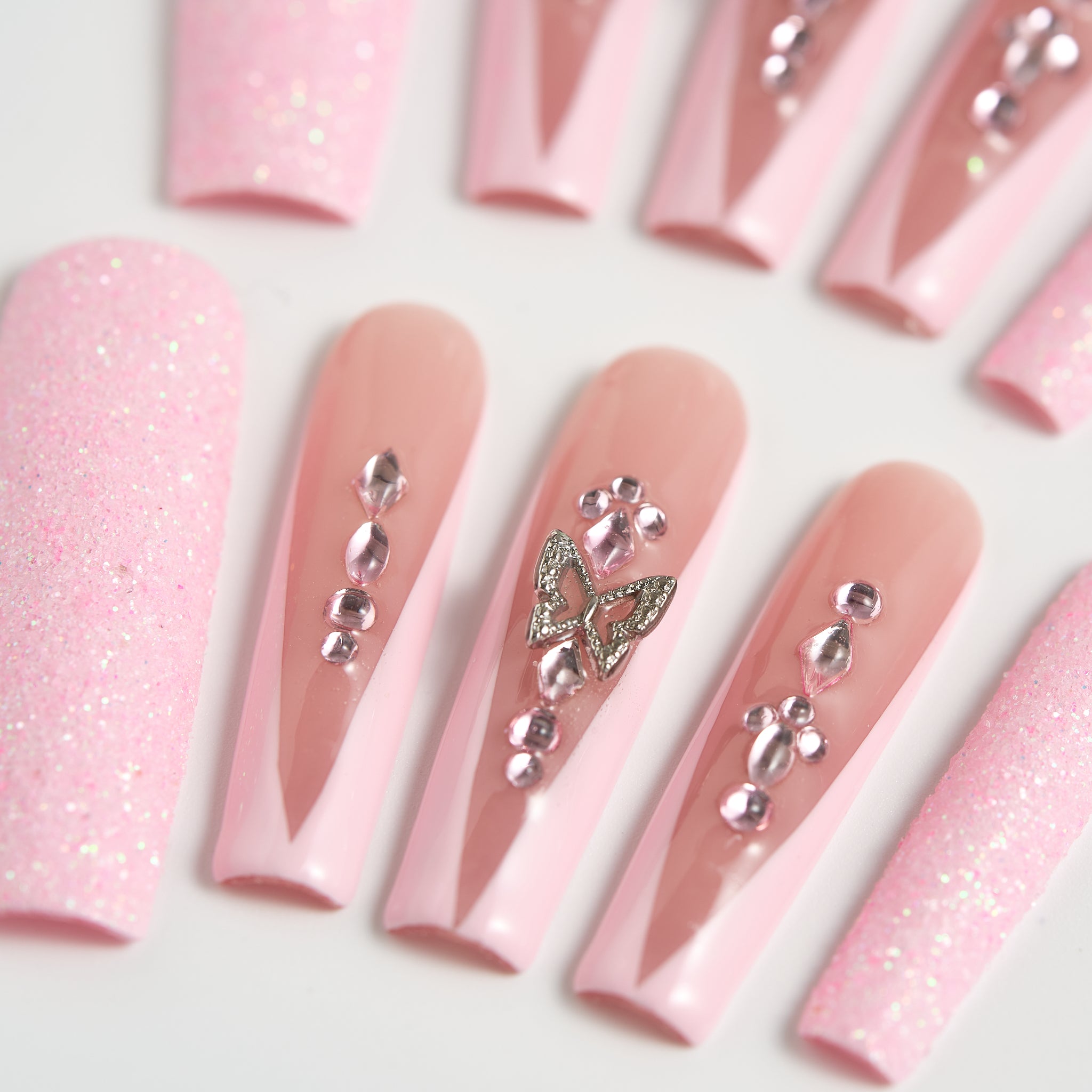 Detailed view of pastel pink and nude press-on nails adorned with clear crystals and a butterfly charm, arranged on a clean white surface.
