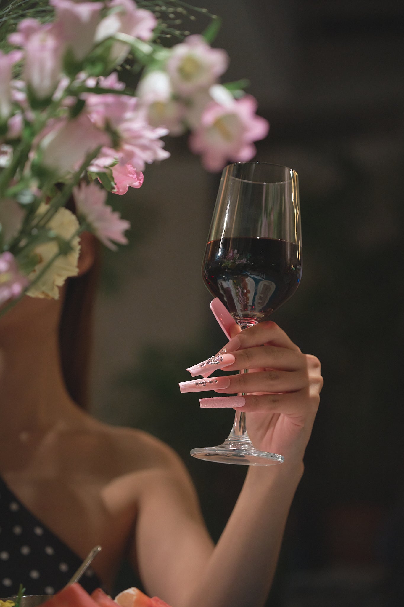 Hand with Sugar Flutter pink press-on nails holding a glass of red wine, blurred floral background adds an elegant touch.