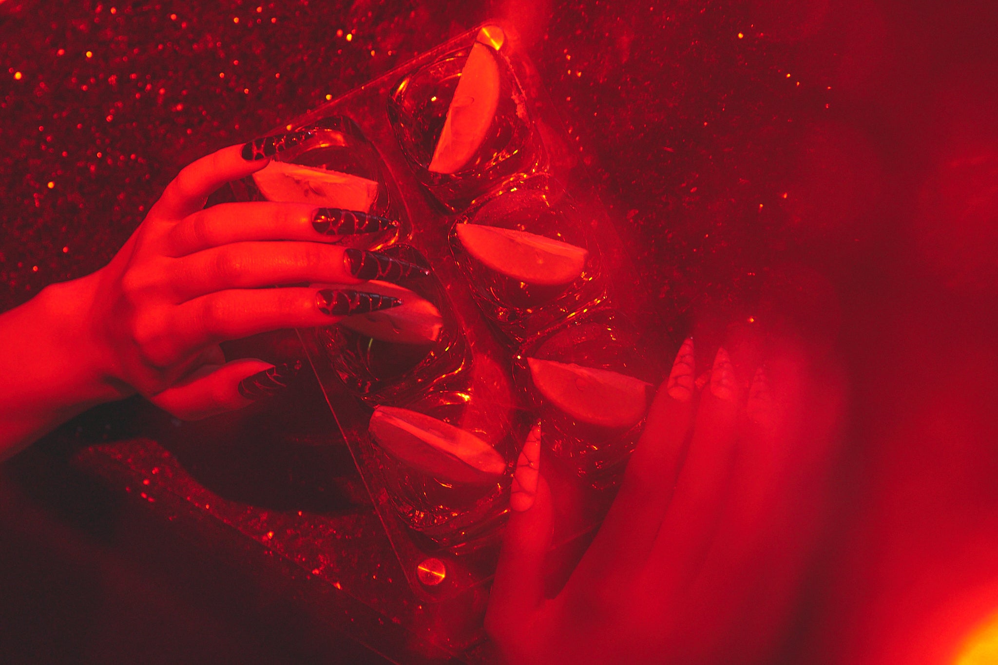 Hands with MiuNail Venom Twist black and white nails holding a cocktail tray under red lighting, surrounded by sliced citrus and shimmering glitter.
