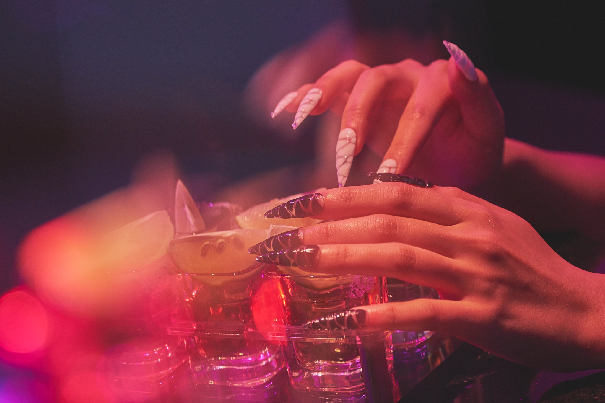 Close-up of hands with MiuNail Venom Twist black and white nails holding a cocktail glass, set against a colorful, blurred nightlife backdrop.