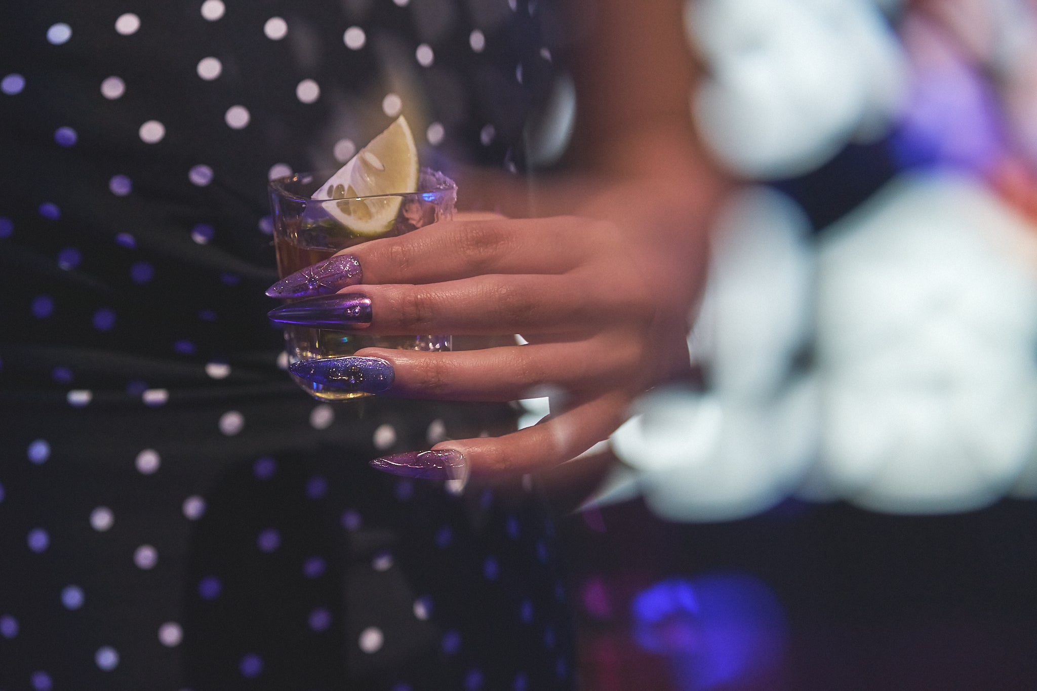 Violet Eclipse stiletto nails in metallic purple and gold, holding a tequila shot glass with a lemon wedge, polka dot dress and nightlife background