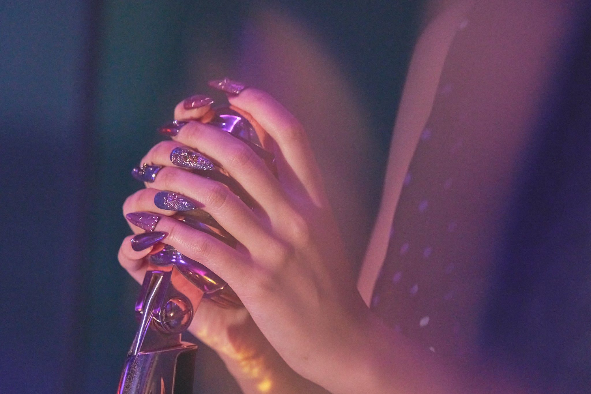 Violet Eclipse stiletto nails in metallic purple and gold, holding a microphone under stage lights