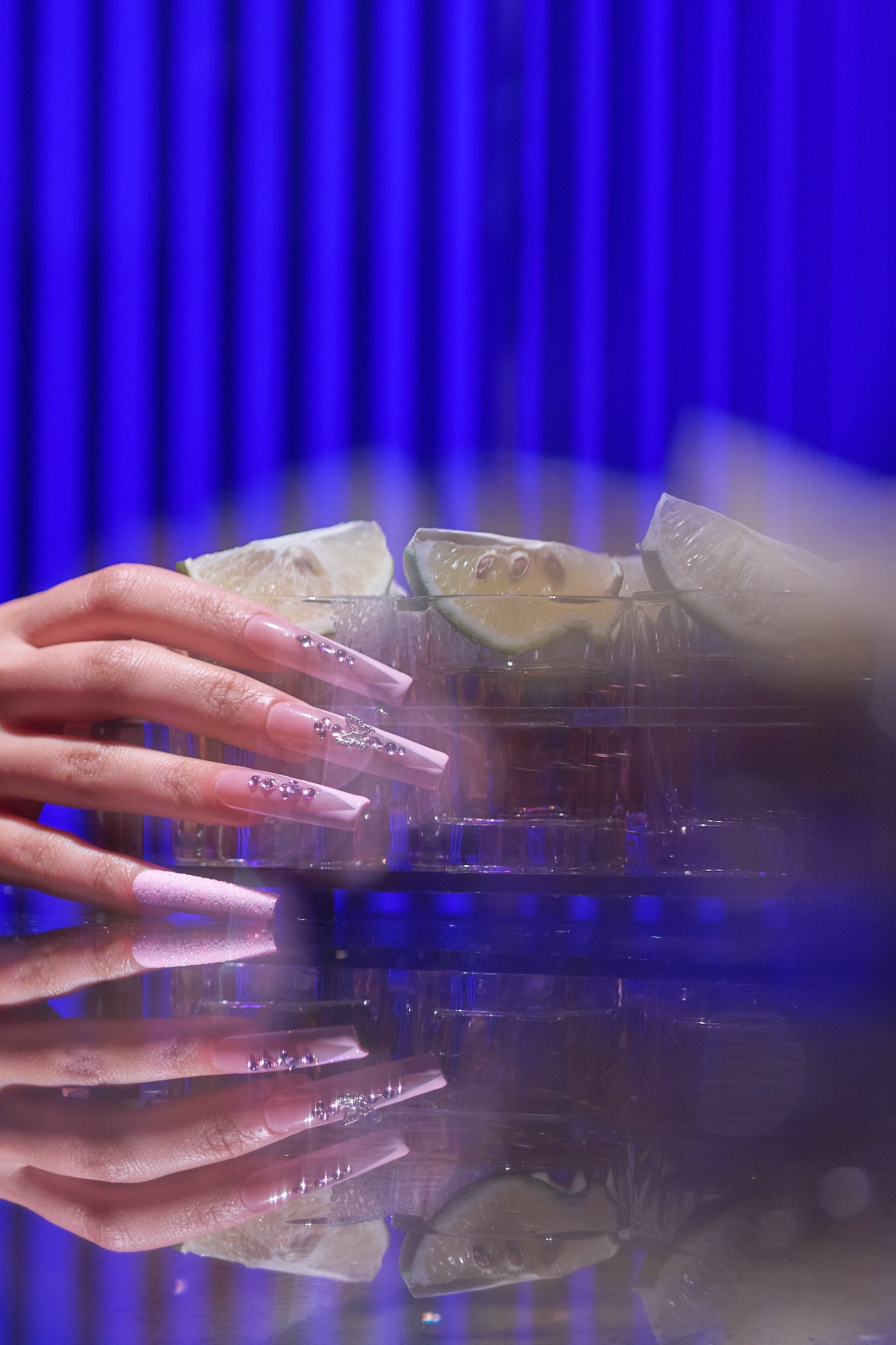 hand with long pink nails holding clear container with lime slices, in front of a vivid blue curtain