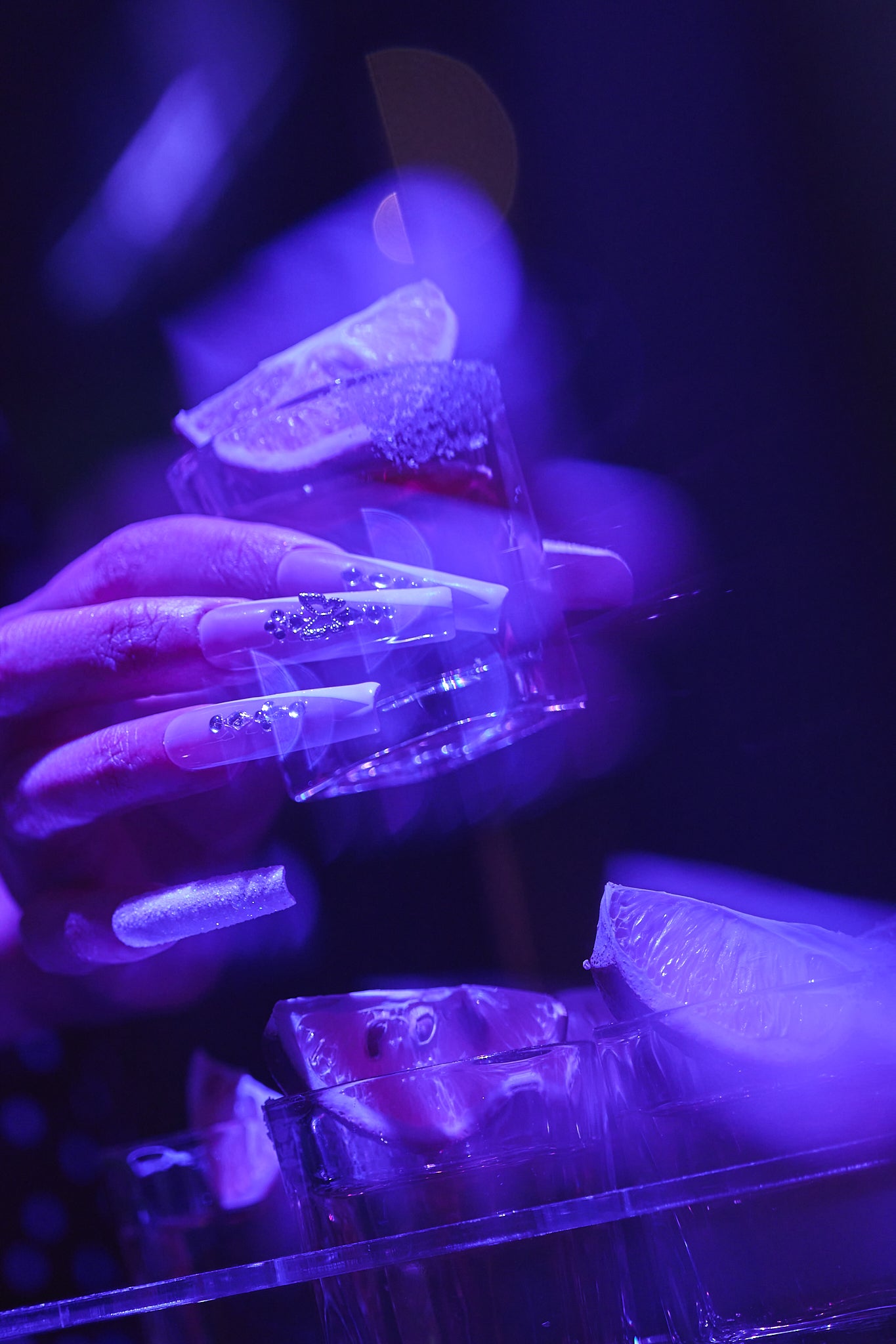 hand with long acrylic nails holding a tilted glass with lime, under violet-blue neon light