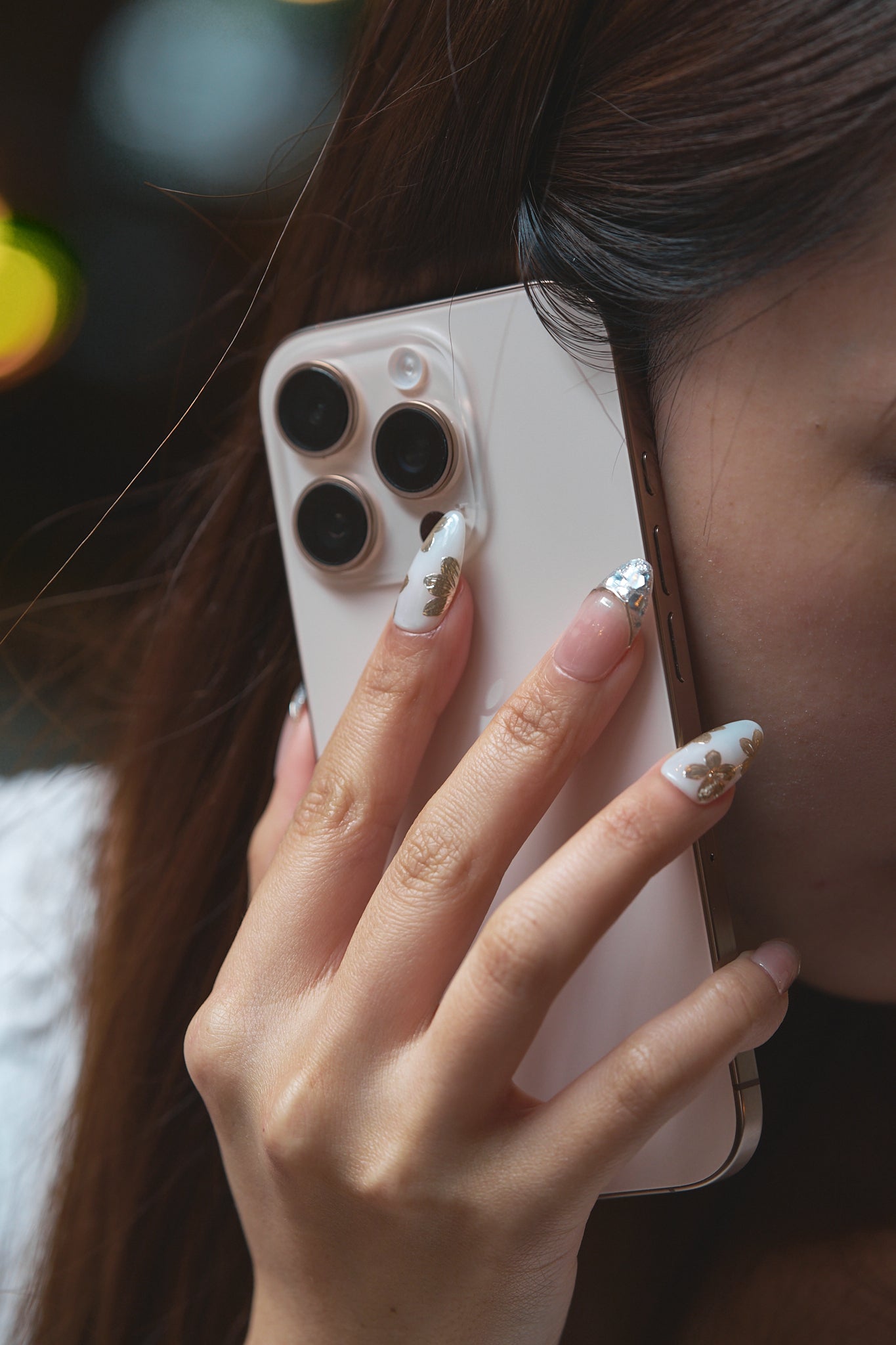 Close-up of a hand with Petal Bloom nails holding a smartphone to the ear, showcasing intricate gold and silver nail designs with a blurred background.