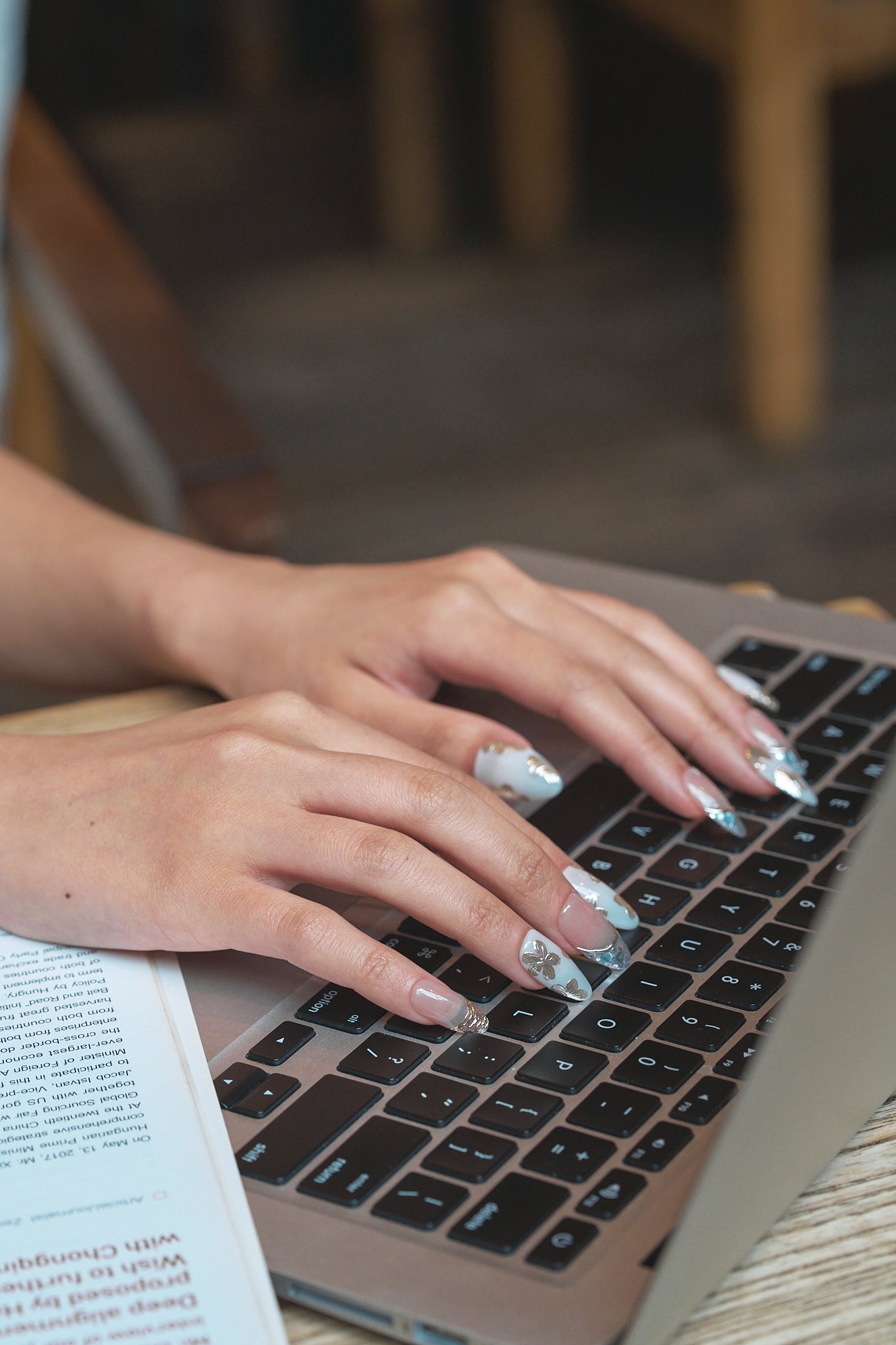 Hands with Petal Bloom decorative nails typing on a laptop keyboard, showing detailed gold and silver nail designs with a blurred background.