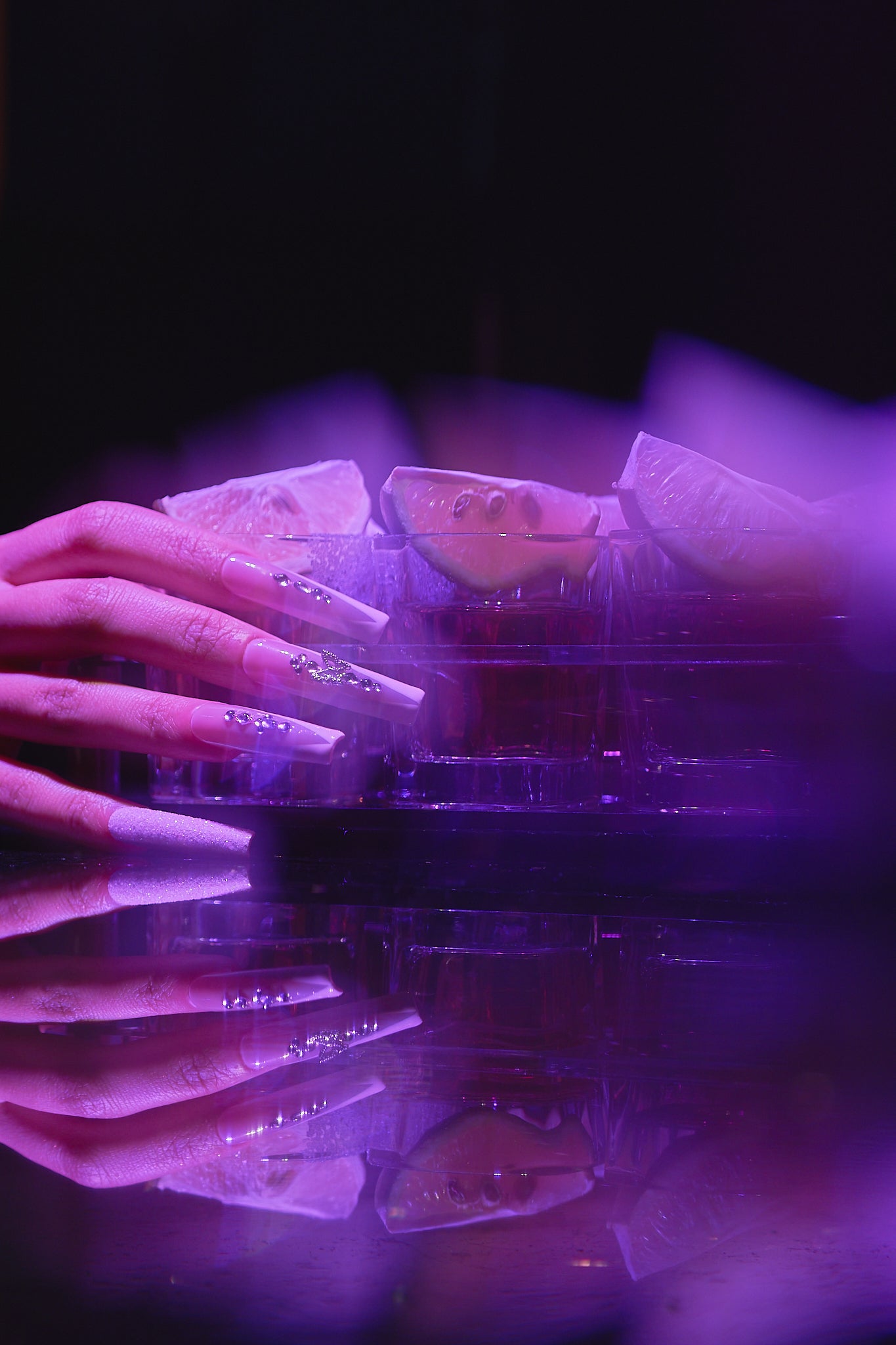 close-up of a hand with long, rhinestone-decorated nails resting on a reflective surface with lemon slices and purple neon lighting