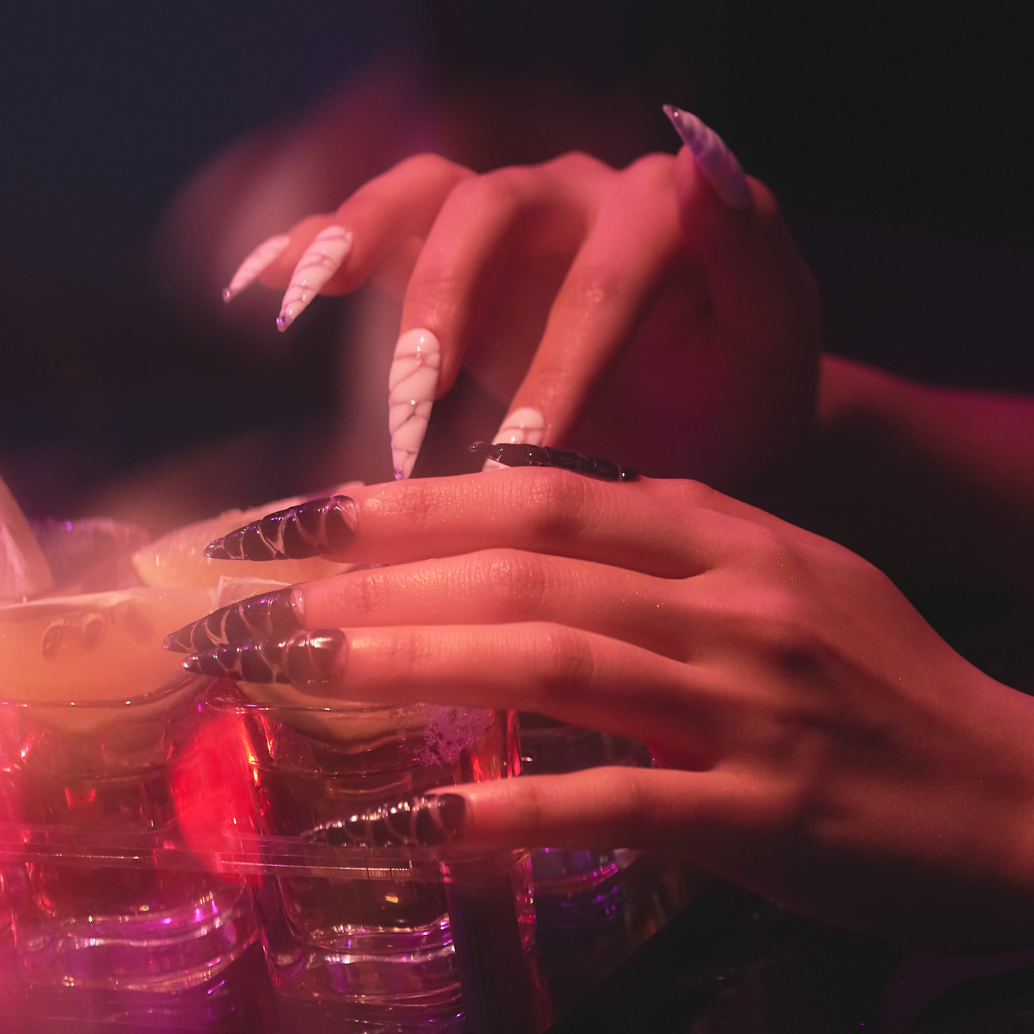 Hand showcasing MiuNail REVERB collection Venom Twist press-on nails with metallic and high-gloss details, holding a textured glass under colorful party lights.